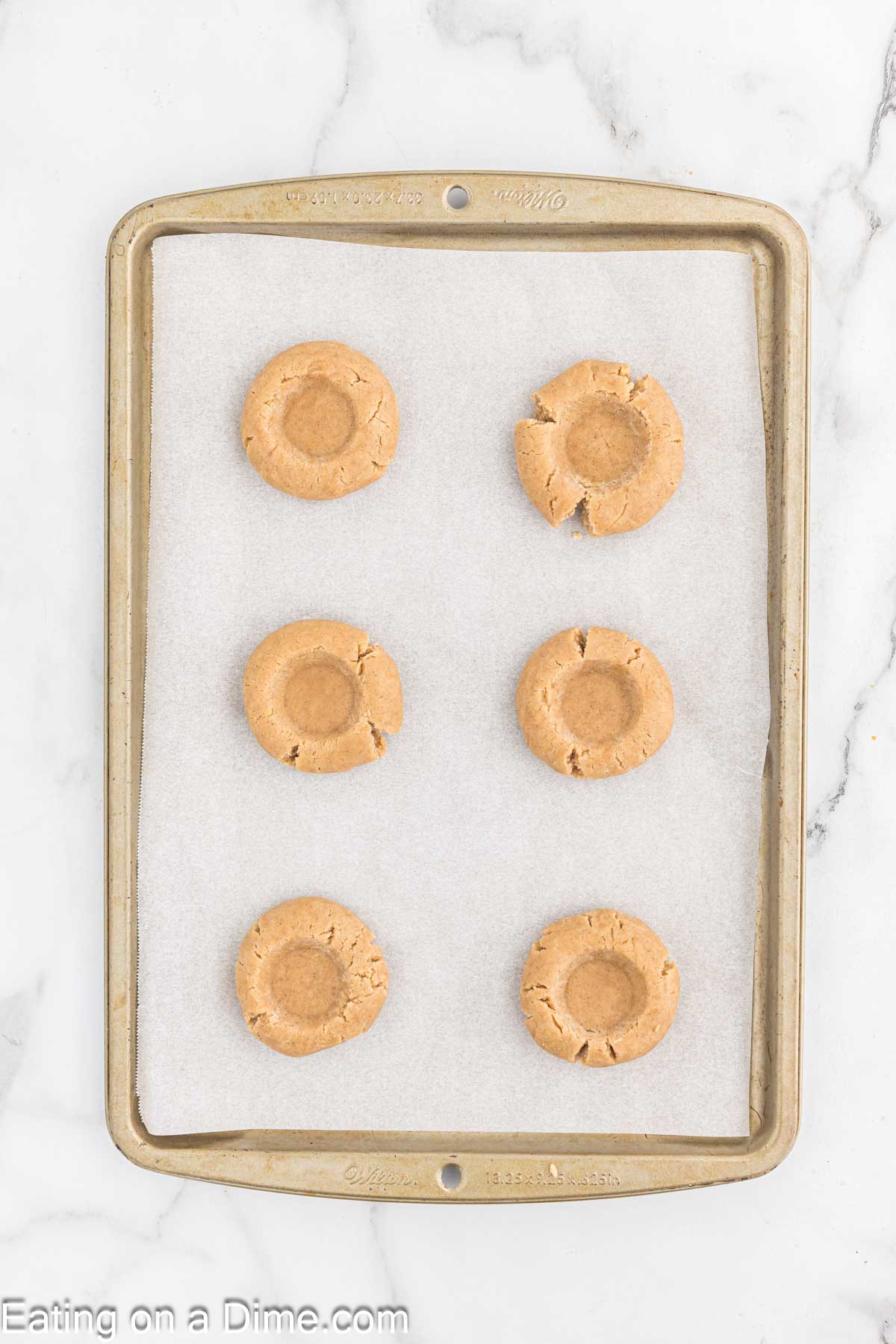 A baking sheet lined with parchment paper holds six evenly spaced peanut butter cookies. Each cookie has a round indentation in the center, perfect for a dollop of jam or a touch of icing. The baking sheet sits on a marble countertop, ready to be paired with an afternoon coffee break.