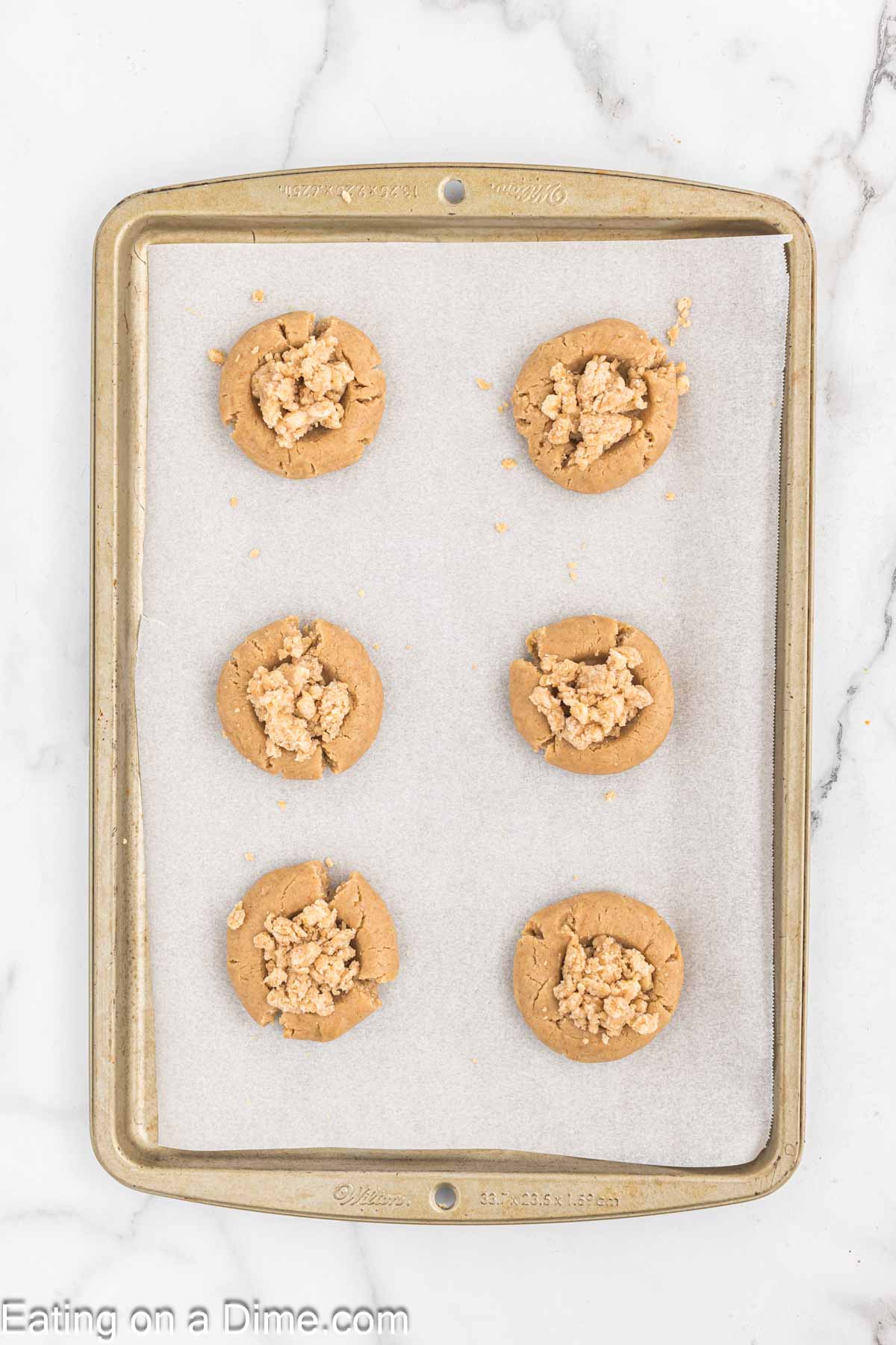 A metal baking tray lined with parchment paper holds six cookies topped with a crumbly, golden brown mixture. The tray rests on a white marble countertop, next to a steaming cup of coffee.