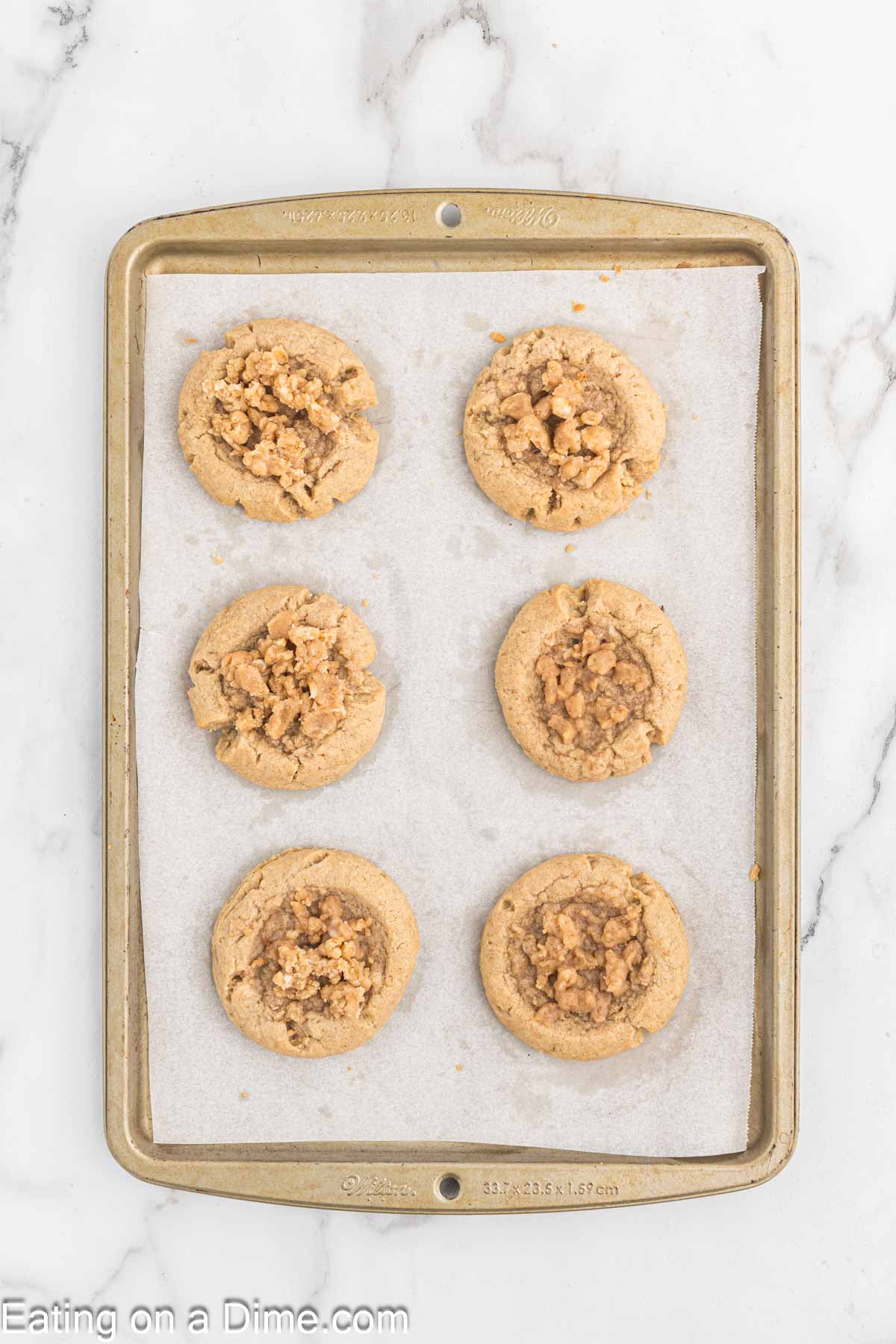 A baking sheet with six evenly spaced cookies topped with a crumbly streusel. The cookies are placed on parchment paper on the baking sheet, which rests on a white marble surface next to a steaming cup of coffee. The visible text on the bottom left says "Eating on a Dime.com.