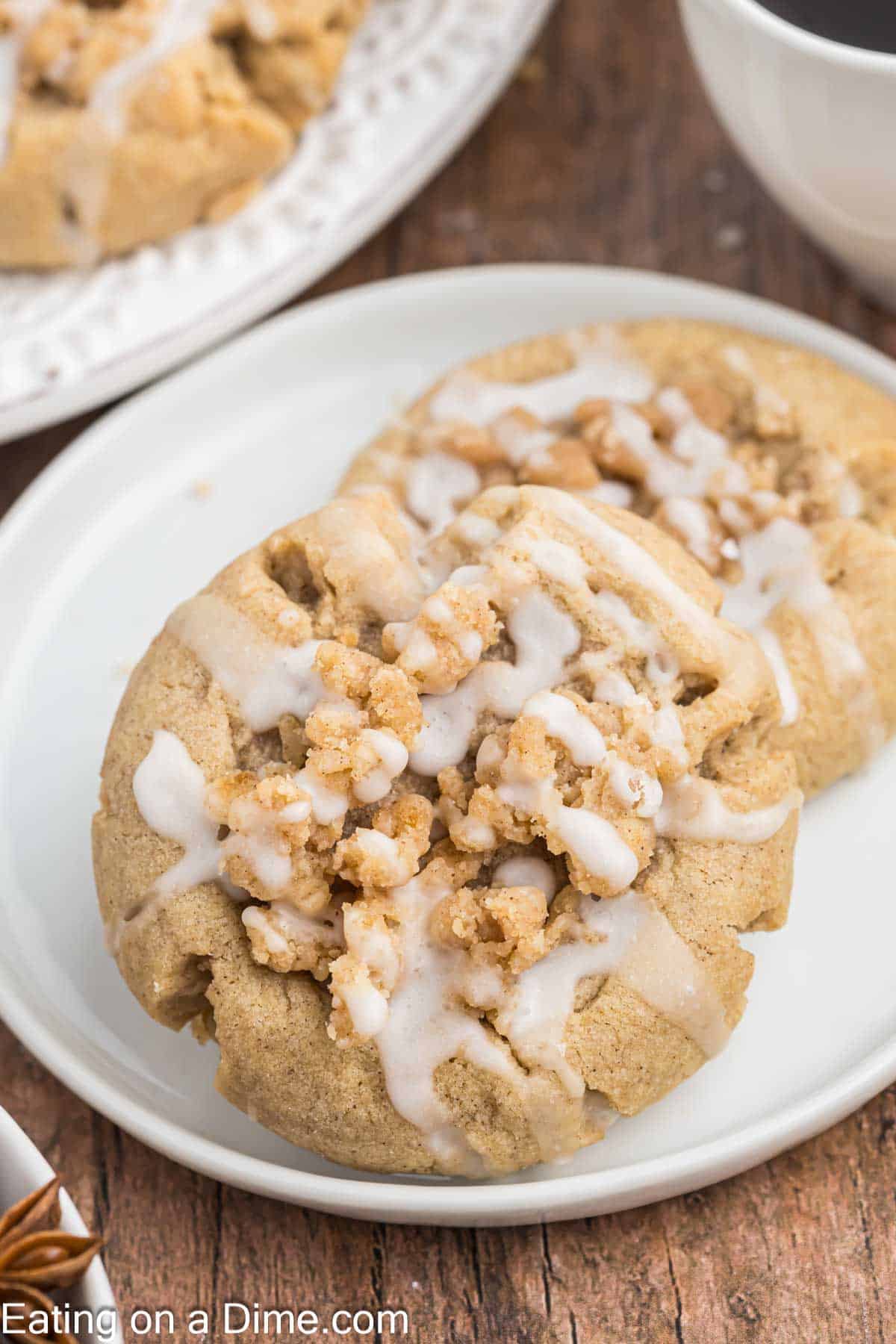 Close-up of two crumbly cookies topped with glaze on a white plate. The cookies have a slightly golden brown color and a generous amount of crumbly topping. A cup of coffee is partially visible in the background on a wooden surface, perfect for pairing with these delectable cookies.