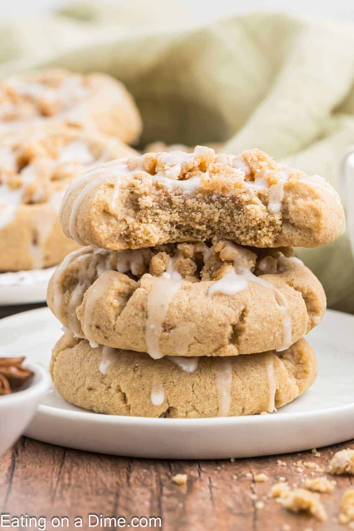 A close-up image of three stacked cookies with a light glaze drizzled on top and crumbled bits. The top cookie has a bite taken out of it. The cookies are on a white plate with more cookies and a green cloth blurred in the background, perfect for pairing with your morning coffee.