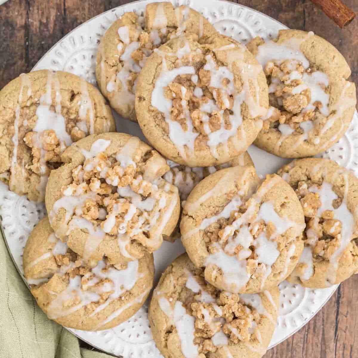 A white decorative plate holds a batch of crinkle cookies topped with a white glaze and sprinkled with a crumbly topping. The cookies, perfect for pairing with coffee, are arranged in a slightly overlapping manner on a wooden surface. A green cloth is partially visible in the bottom left corner.