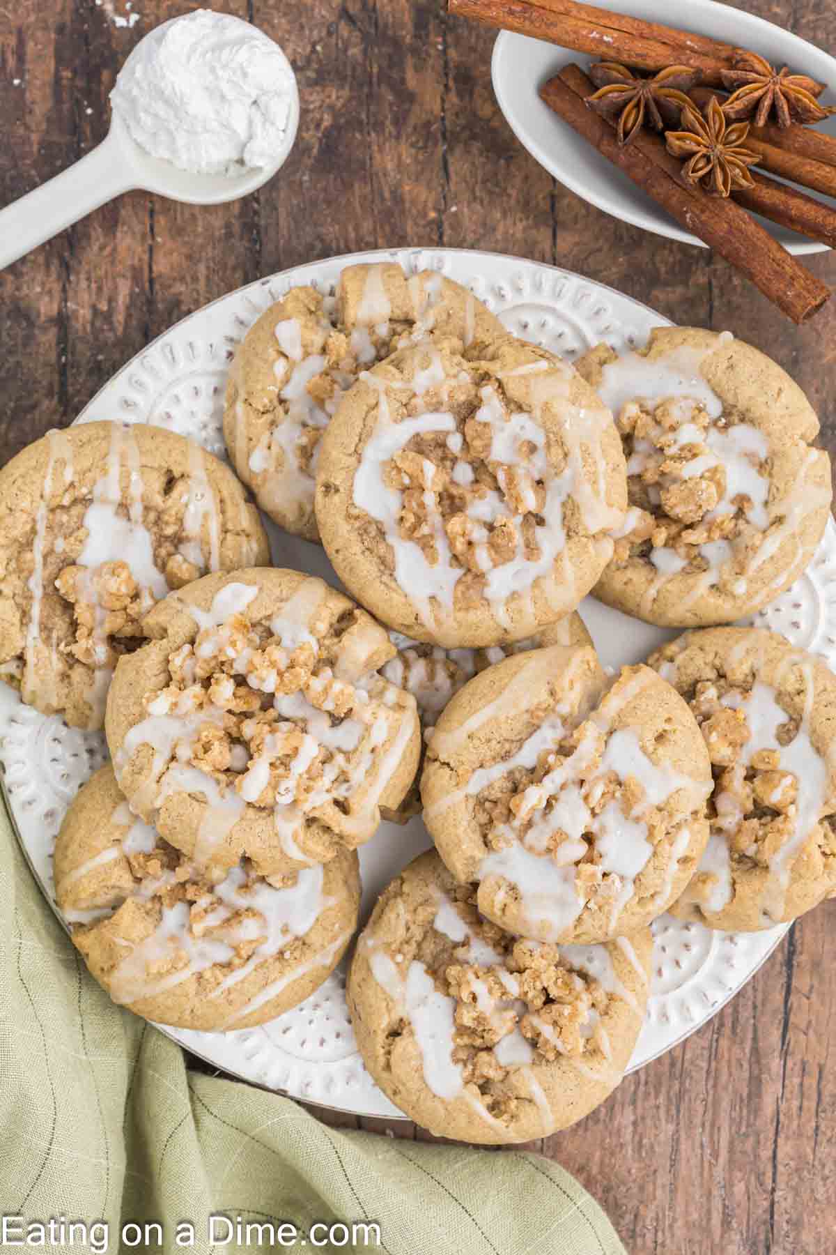 A plate of nine iced coffee cake cookies rests on a white, intricately designed plate placed on a wooden surface. A bowl of powdered sugar and scattered cinnamon sticks create a charming background, while a light green napkin is partially visible in the foreground.