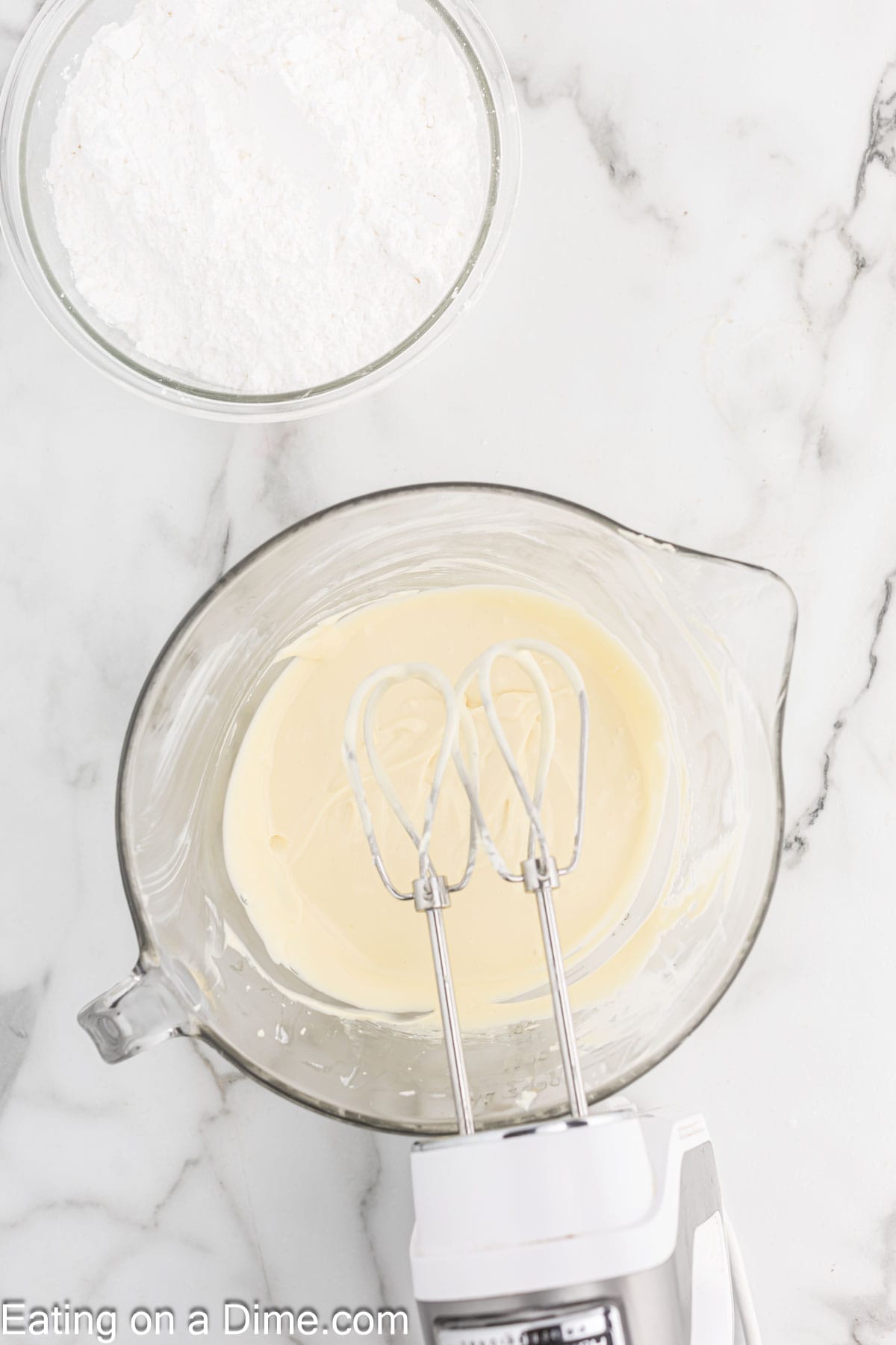 Cream cheese mixture in a bowl with a hand mixer with a bowl of powdered sugar