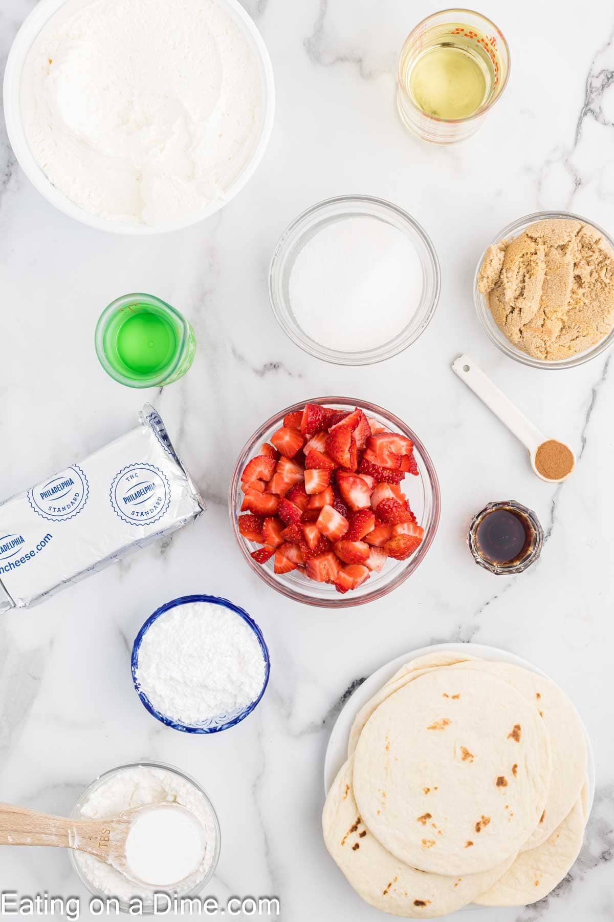 An assortment of ingredients on a marble countertop, including a plate of tortillas, a bowl of diced strawberries, brown sugar, cream cheese, powdered sugar, cinnamon, vanilla extract, cooking oil, green and clear liquids, and white sugar. Perfect for crafting dreamy strawberry cheesecake tacos.