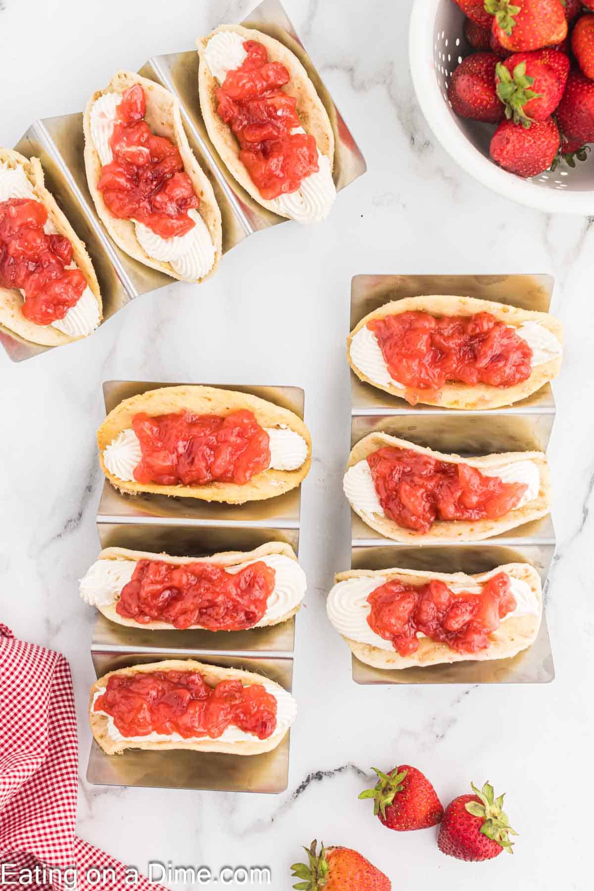 Eight dessert tacos filled with cheesecake cream and topped with strawberry compote are arranged on a marble surface. A bowl of fresh strawberries sits in the top right corner, and a red and white checkered cloth is partially visible in the bottom left corner.