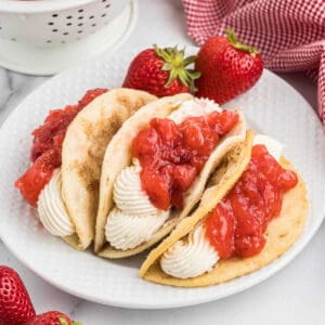 A white plate holds three dessert tacos filled with piped whipped cream and topped with chunky strawberry sauce, reminiscent of a delightful cheesecake. Fresh strawberries and a red and white checkered cloth are in the background, along with a white colander.