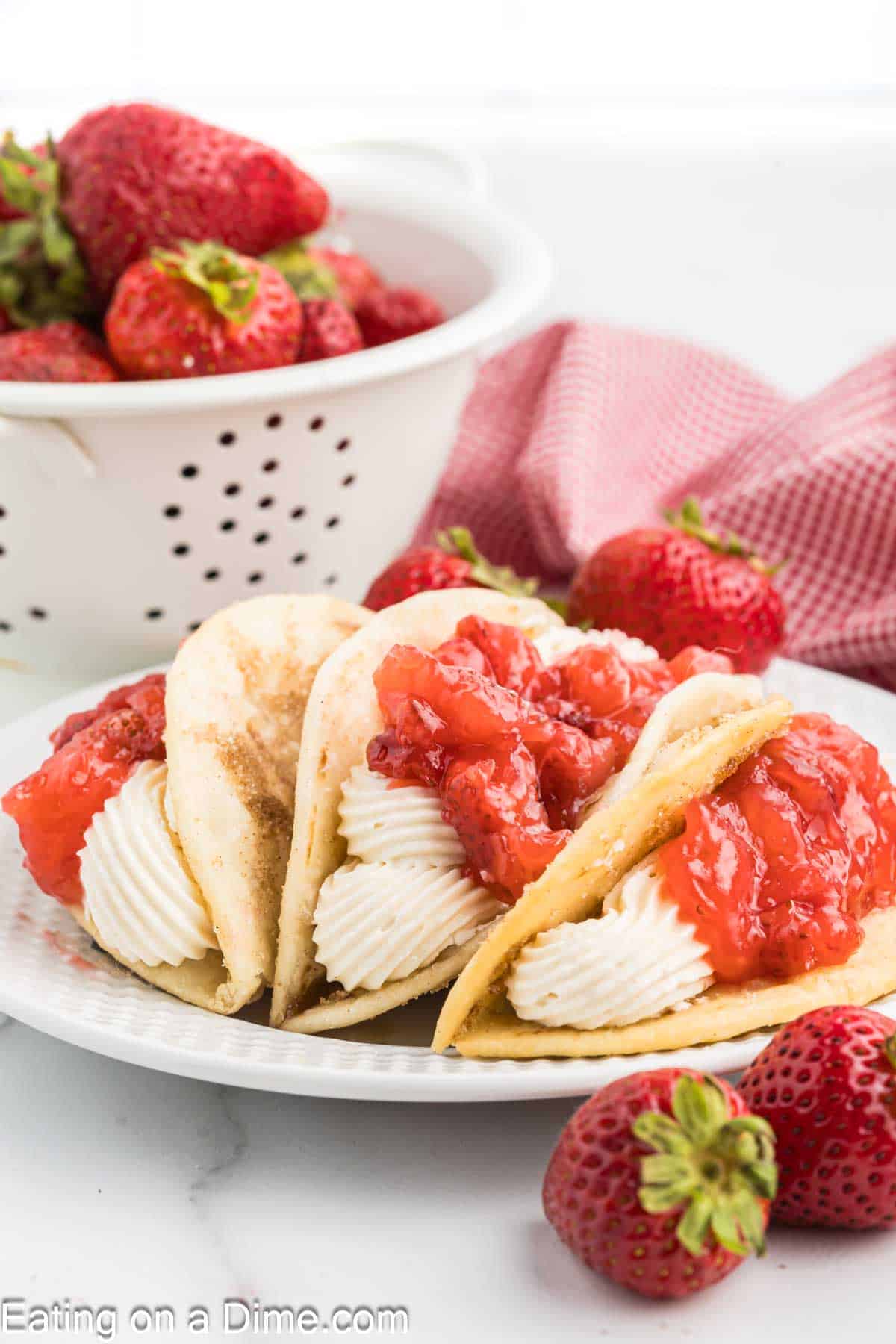 A plate with three Cheesecake Tacos is shown. The taco-shaped desserts are filled with whipped cream and topped with a chunky strawberry sauce. Fresh strawberries are scattered alongside, and a white colander filled with strawberries and a red gingham napkin are in the background.