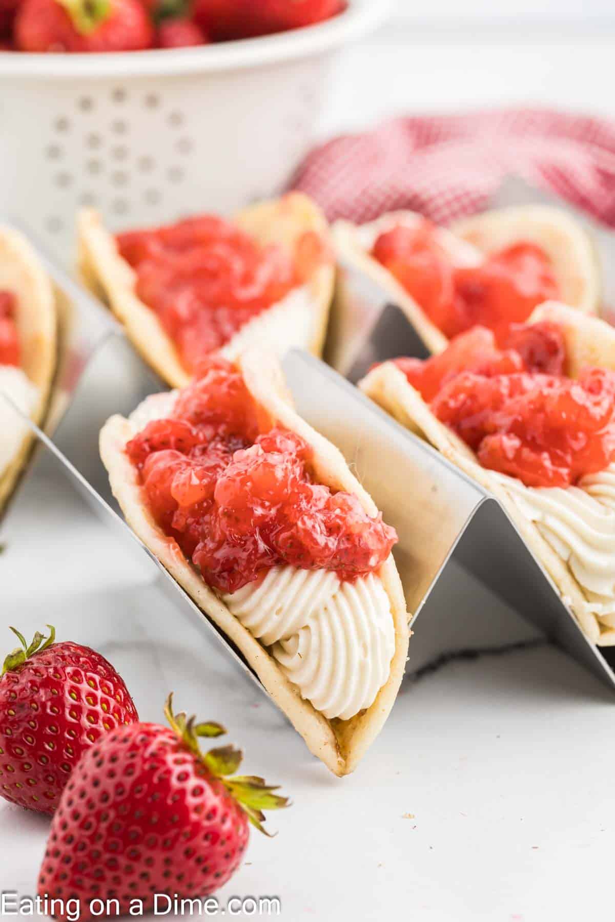 Cheesecake Tacos filled with cream cheese frosting and topped with chunky strawberry sauce are arranged in metal taco holders. Fresh strawberries and a red checkered cloth in the background add a vibrant and fresh touch to the image.