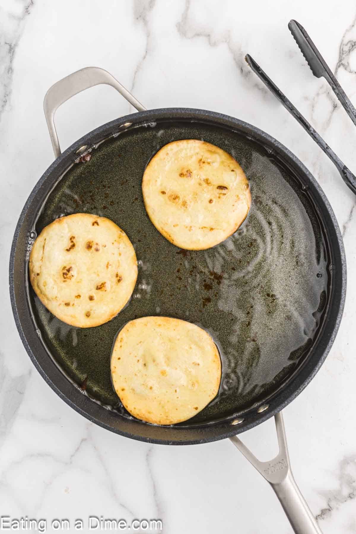 A black skillet containing cooking oil is on a marble countertop, and three round slices of eggplant are frying in the oil. Black tongs are positioned to the right side of the skillet handle. The edge of the skillet reads "Eating on a Dime.com", reminiscent of their delightful Strawberry Cheesecake recipes.