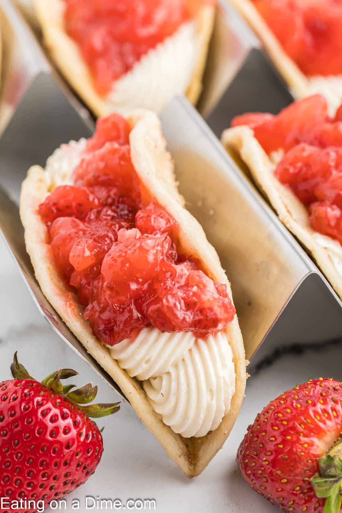 Close-up of strawberry shortcake tacos, featuring crispy taco shells filled with piped white cream and topped with chunks of fresh strawberries. Two whole strawberries are in the foreground, adding a fresh touch to this cheesecake-inspired dessert display.