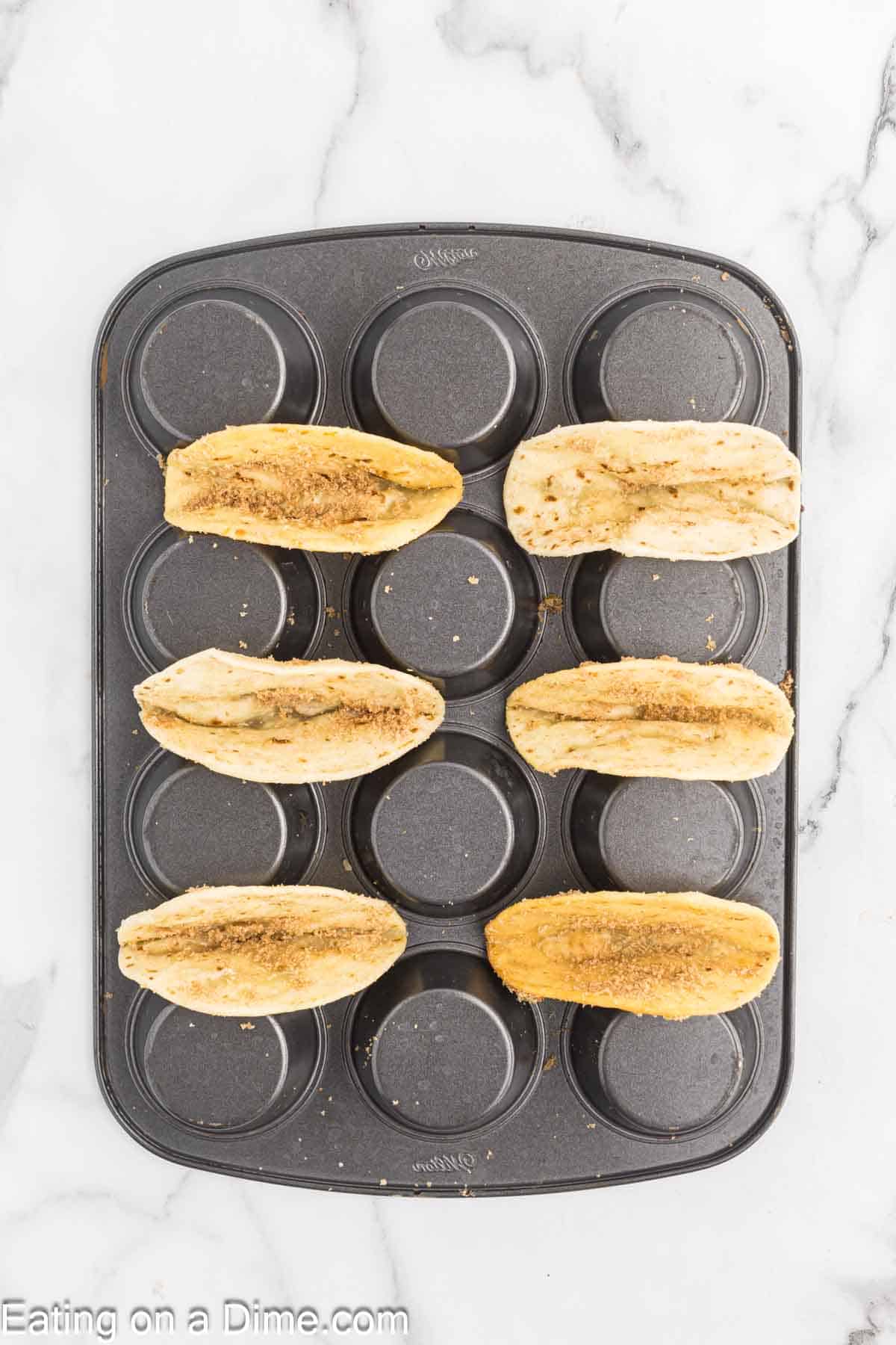 A muffin pan is shown with six hot dog buns placed in the cavities. The buns are seasoned, cut-side up, and toasted. The pan is situated on a white marble countertop next to a tantalizing strawberry cheesecake.