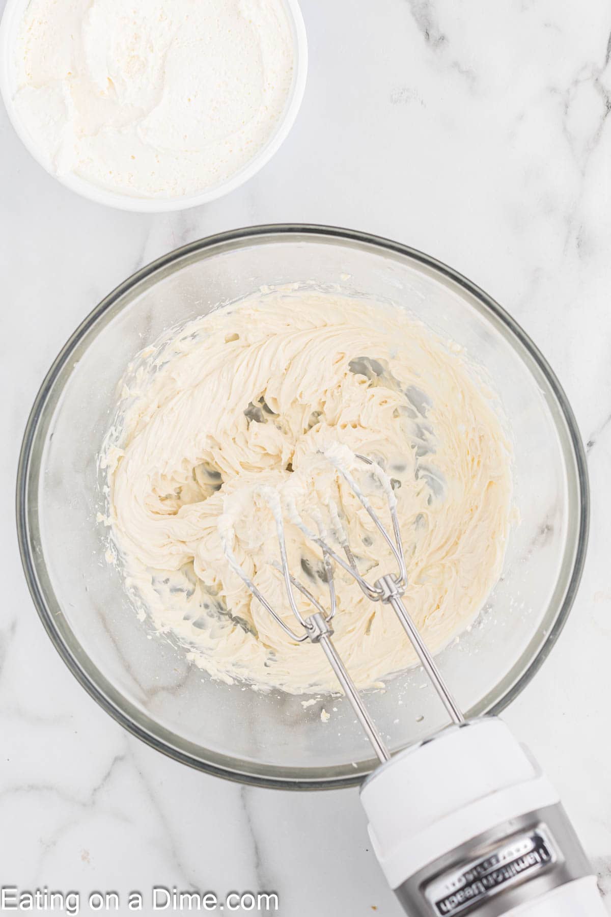 A mixing bowl filled with creamy whipped mixture is shown on a marble countertop. An electric hand mixer with beaters is partially immersed in the bowl, perfect for making Strawberry Cheesecake Tacos. Above the bowl, there's a container with more of the same mixture. Text at the bottom reads "Eating on a Dime.com.