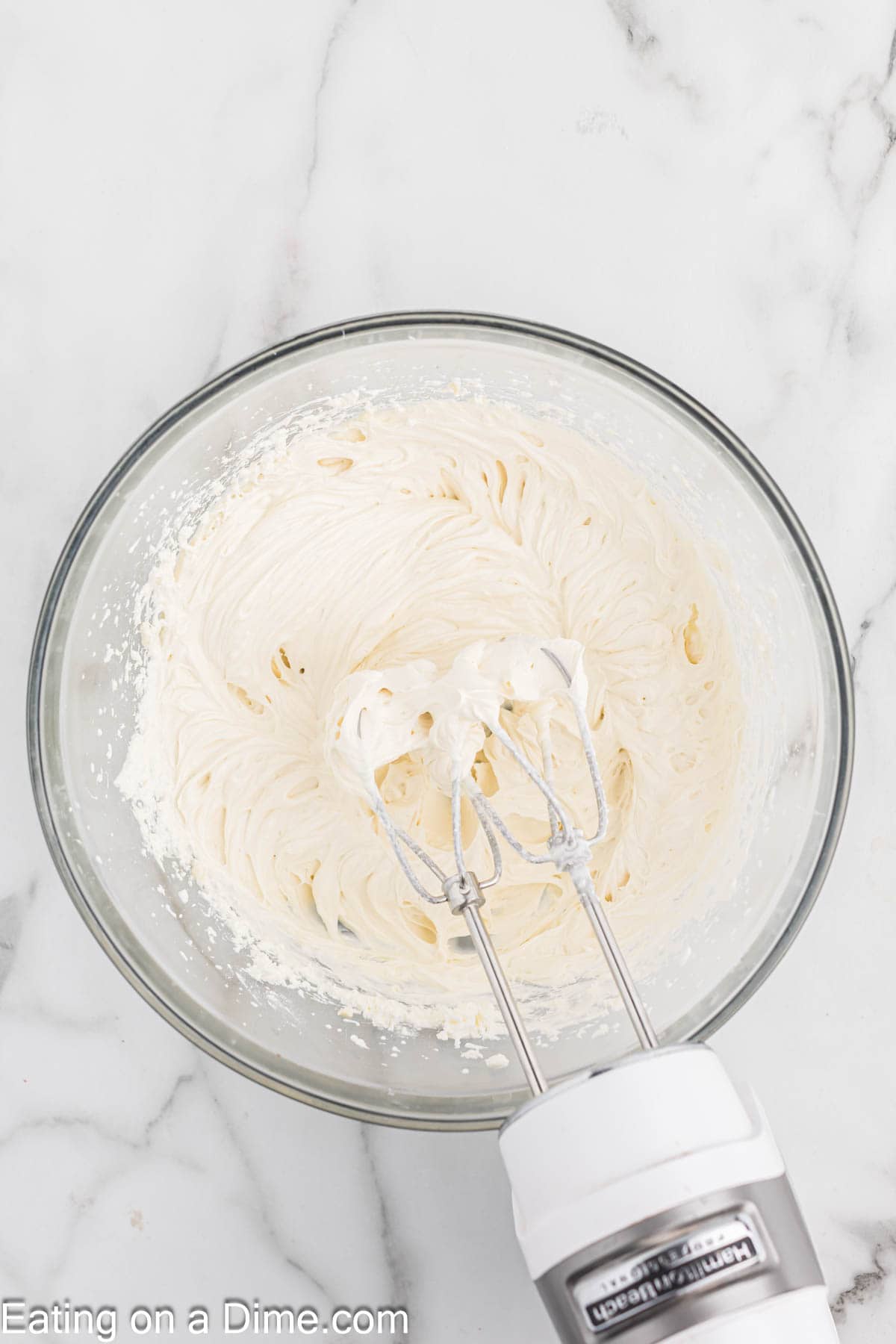 A glass bowl filled with smooth, creamy white frosting being mixed with an electric hand mixer on a marble countertop. The beaters are partially immersed in the frosting, and a small amount of frosting clings to them. Imagine this luscious icing atop your favorite cheesecake. The text "Eating on a Dime.com" is at the bottom.