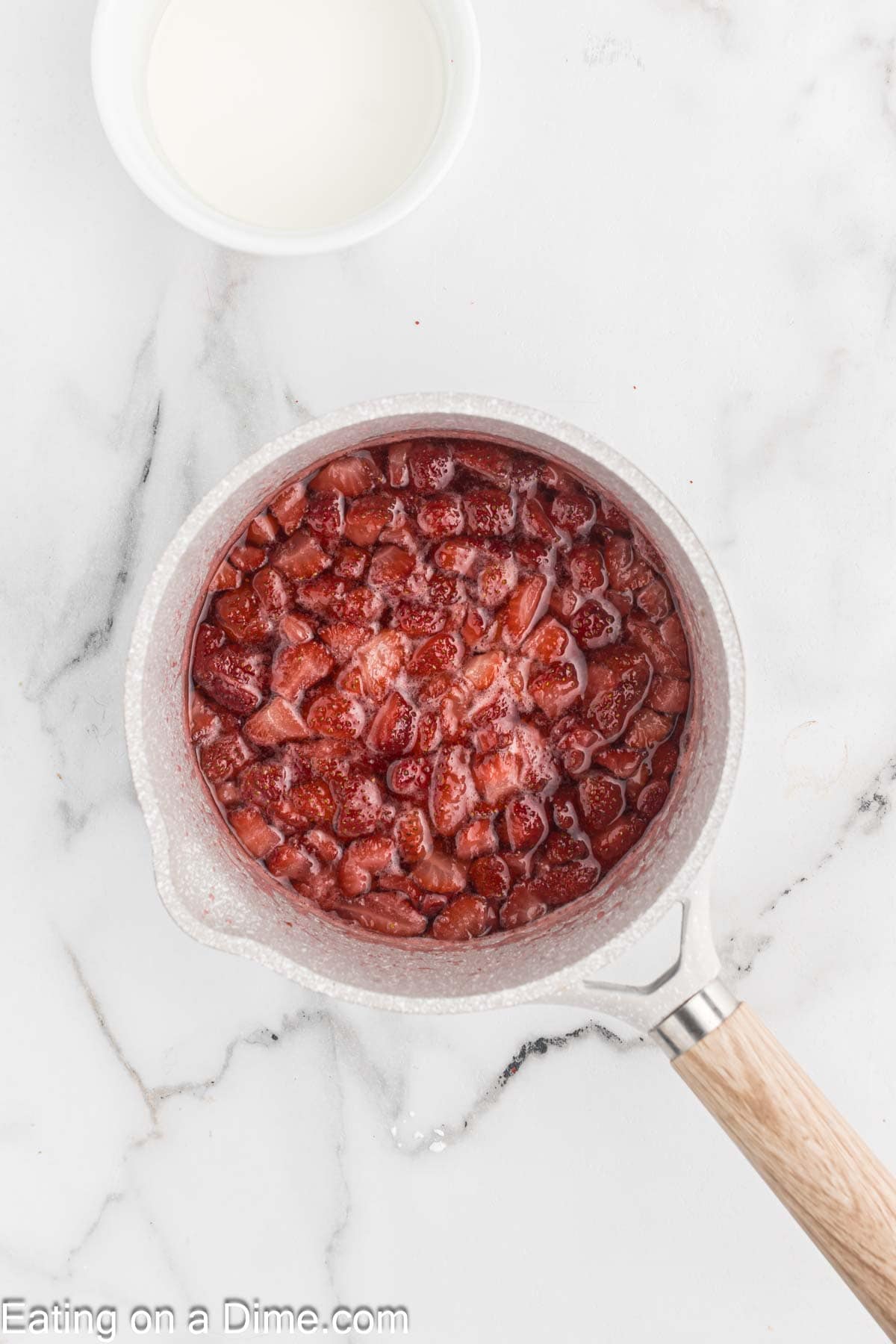 A white saucepan filled with chopped strawberries simmering in a red liquid is placed on a white marble countertop, hinting at delicious Strawberry Cheesecake Tacos. Above the saucepan, there is a small white bowl containing milk. The text "Eating on a Dime.com" appears in the bottom left corner.