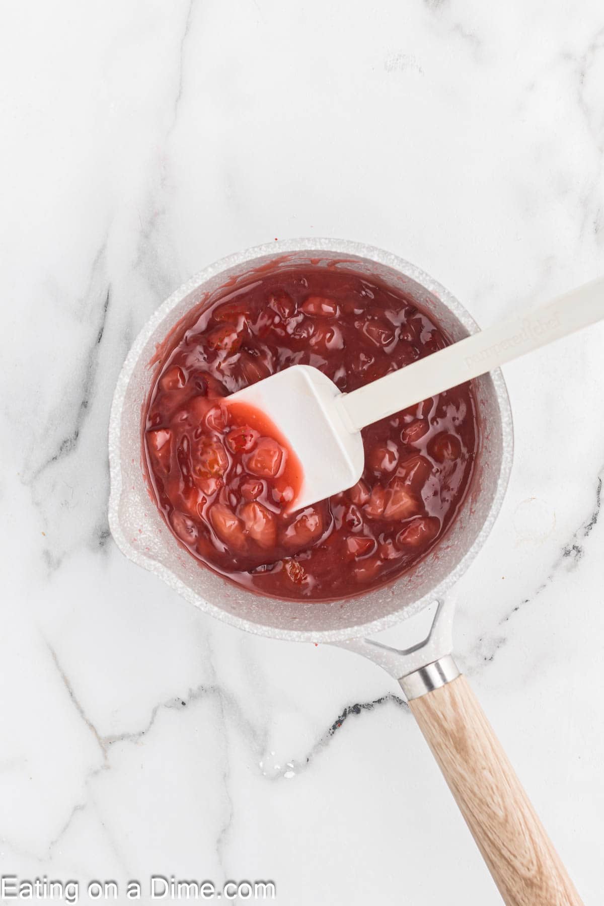 A white saucepan filled with a chunky red sauce sits on a marble countertop. The sauce, looking like it contains pieces of strawberry, embraces a white spatula with a wooden handle partially dipped in. The words "Eating on a Dime.com" are partially visible at the bottom of the image.