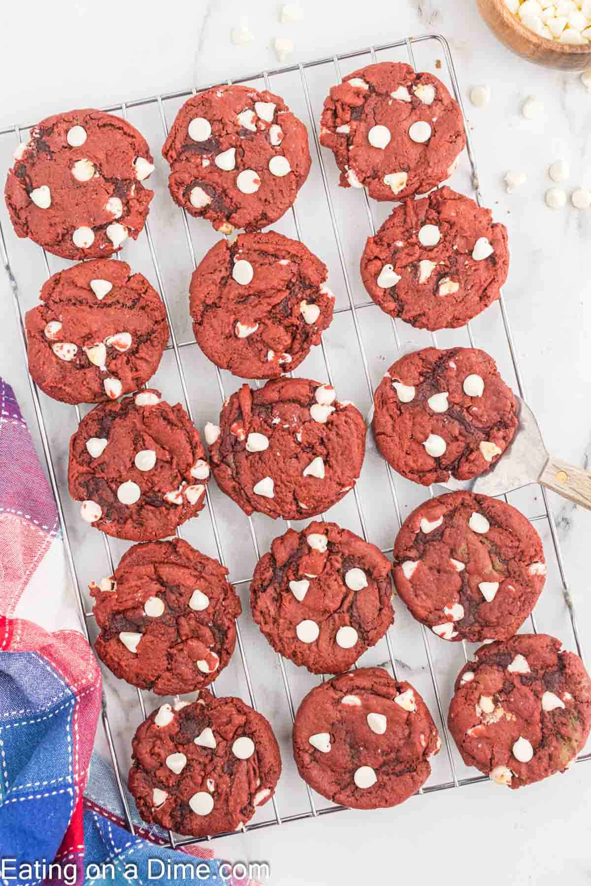 A cooling rack filled with red velvet cookies topped with white chocolate chips sits on a marble countertop. A colorful cloth is partially visible in the bottom left, and a bowl of extra white chocolate chips is in the top right corner.