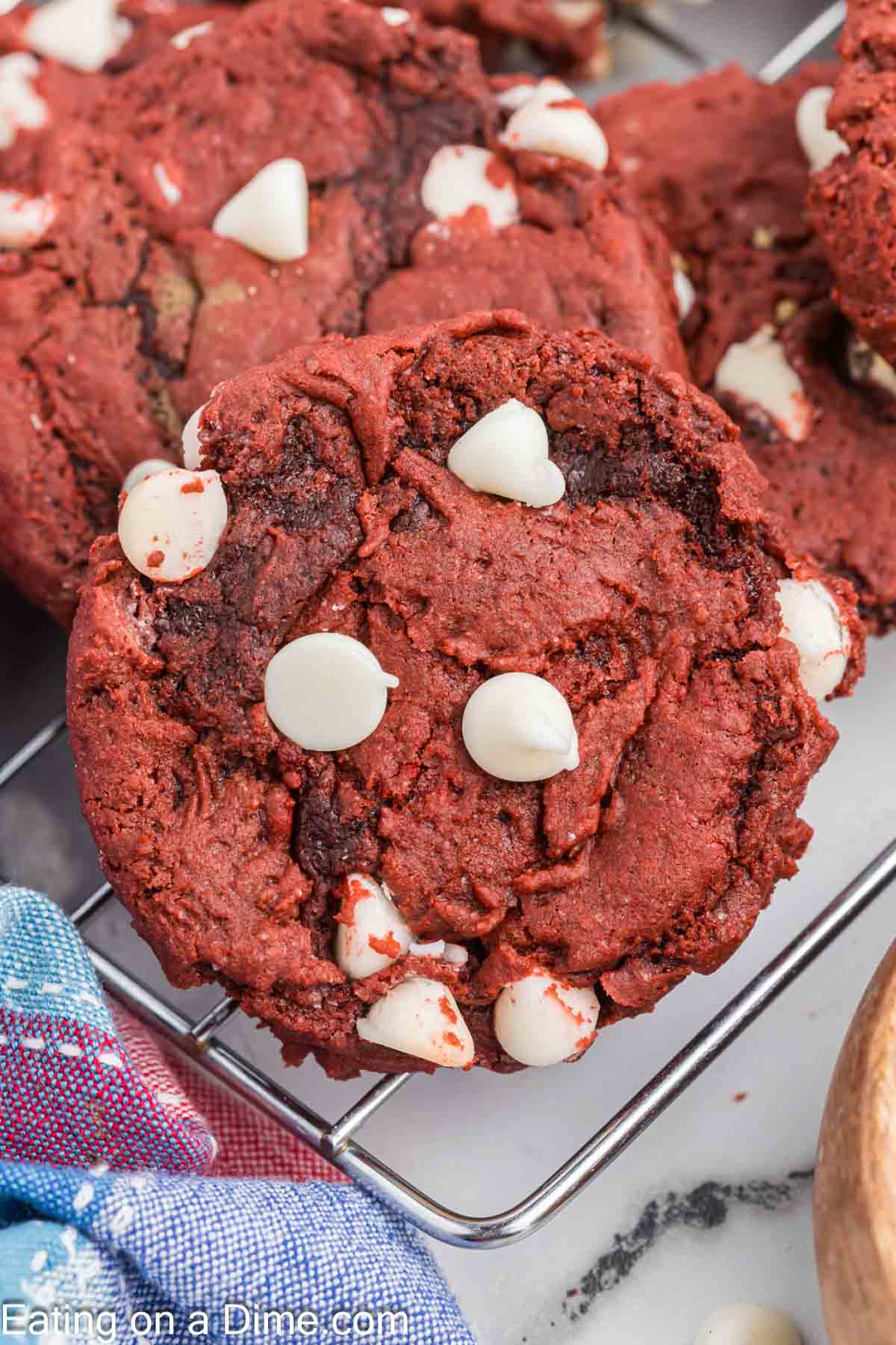 Close-up of a cooling rack holding freshly baked Red Velvet cookies studded with white chocolate chips. The cookies have a rich red hue and are slightly cracked on the surface. A colorful cloth is partially visible in the bottom left corner.