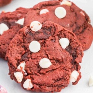 Three red velvet cookies are displayed, topped with several white chocolate chips. The cookies have a slightly cracked surface and rich red color, with the white chocolate chips providing a stark contrast. A pink-tinged cloth is partially visible in the corner, enhancing the vibrant scene of these delightful red velvet cookies.