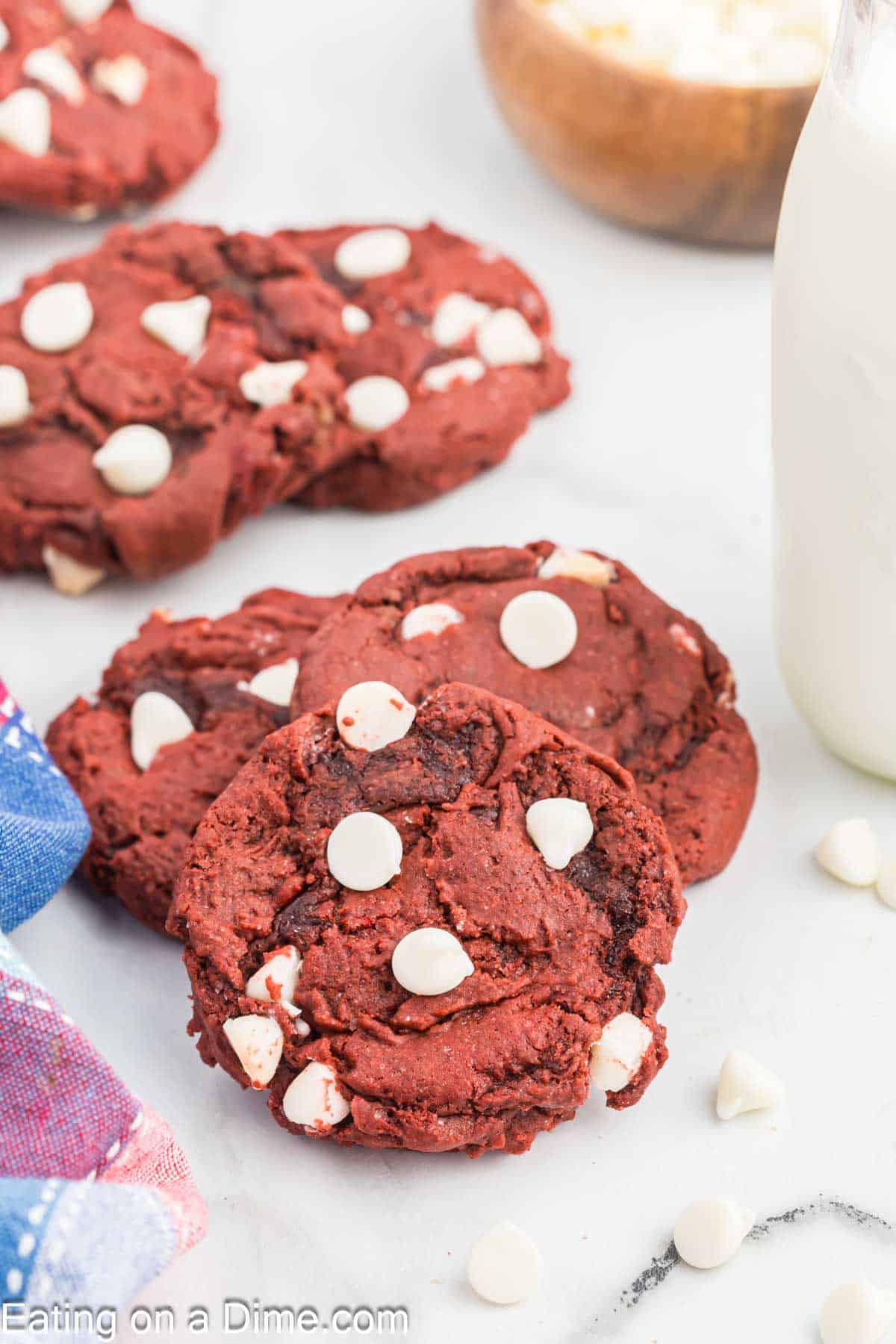 Three red velvet cookies with white chocolate chips are stacked on each other in the foreground, while more cookies are scattered in the background. A glass of milk sits to the right, and a colorful cloth is partially visible in the lower left corner.