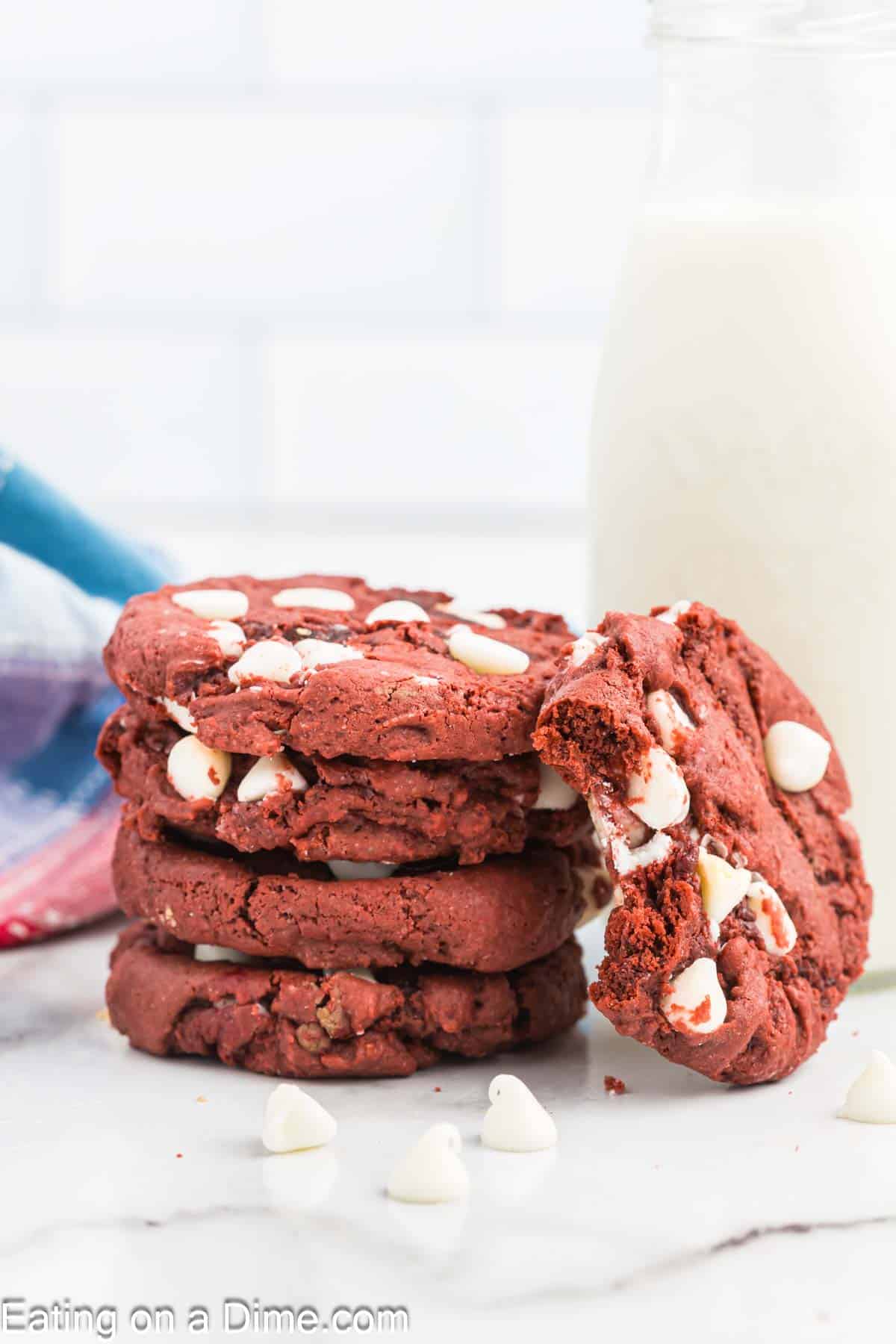 A stack of red velvet cookies with white chocolate chips sits on a white surface. One cookie, with a bite taken out of it, leans against the stack. In the background, there's a partially visible glass bottle of milk and a colorful cloth.