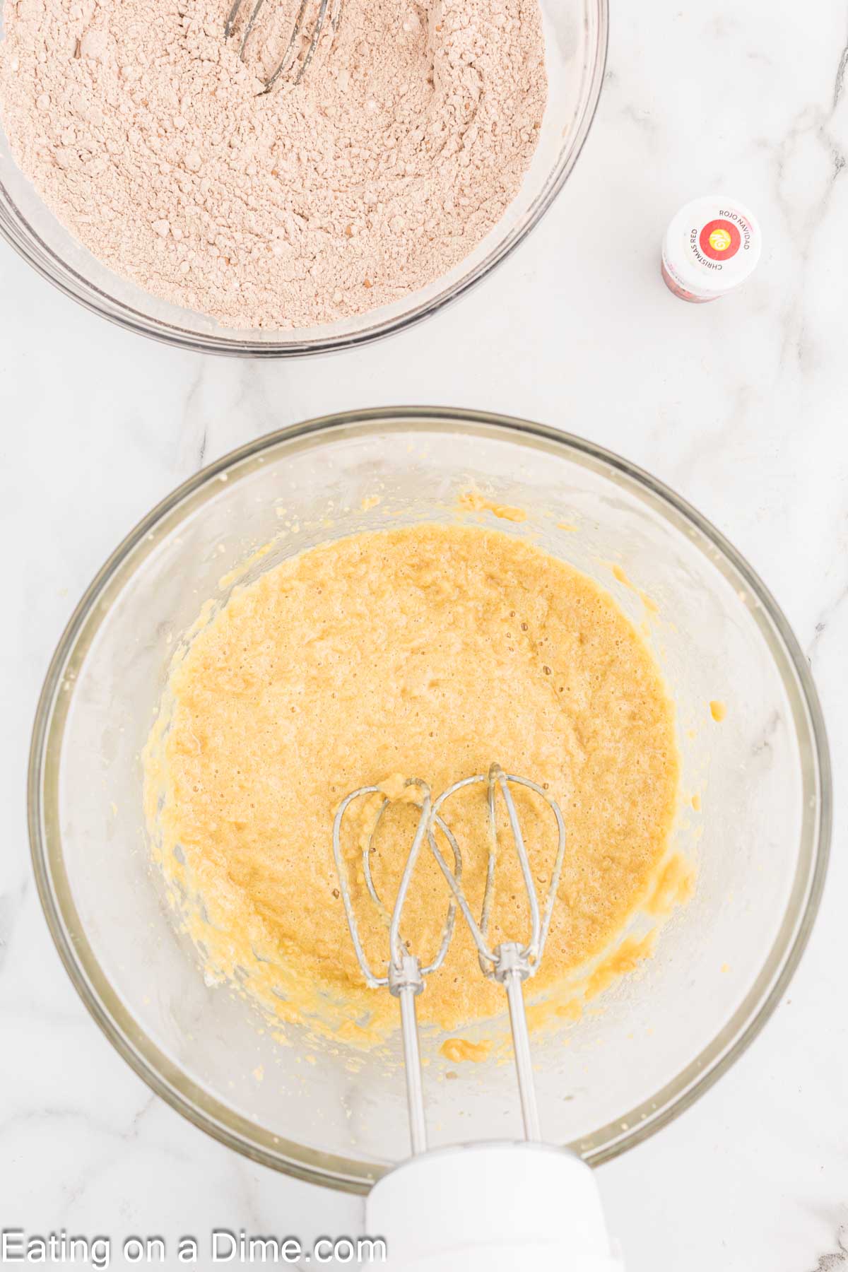 Two mixing bowls: one with a whisk and brown dry ingredients in the top left, and the other with an electric mixer and yellow mixture in the center. A small bottle of red food coloring for Red Velvet Cookies is nearby on the right. The text "Eating on a Dime.com" is at the bottom.