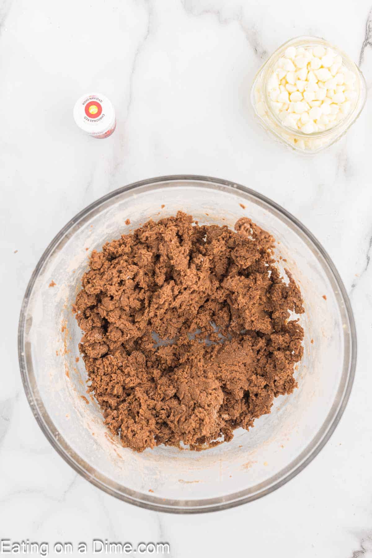 A clear glass bowl filled with cookie dough mixture sits on a marble countertop. Nearby, a small clear bowl contains white chocolate chips, and a small red-capped bottle, possibly food coloring or extract to make Red Velvet Cookies, is placed beside it.