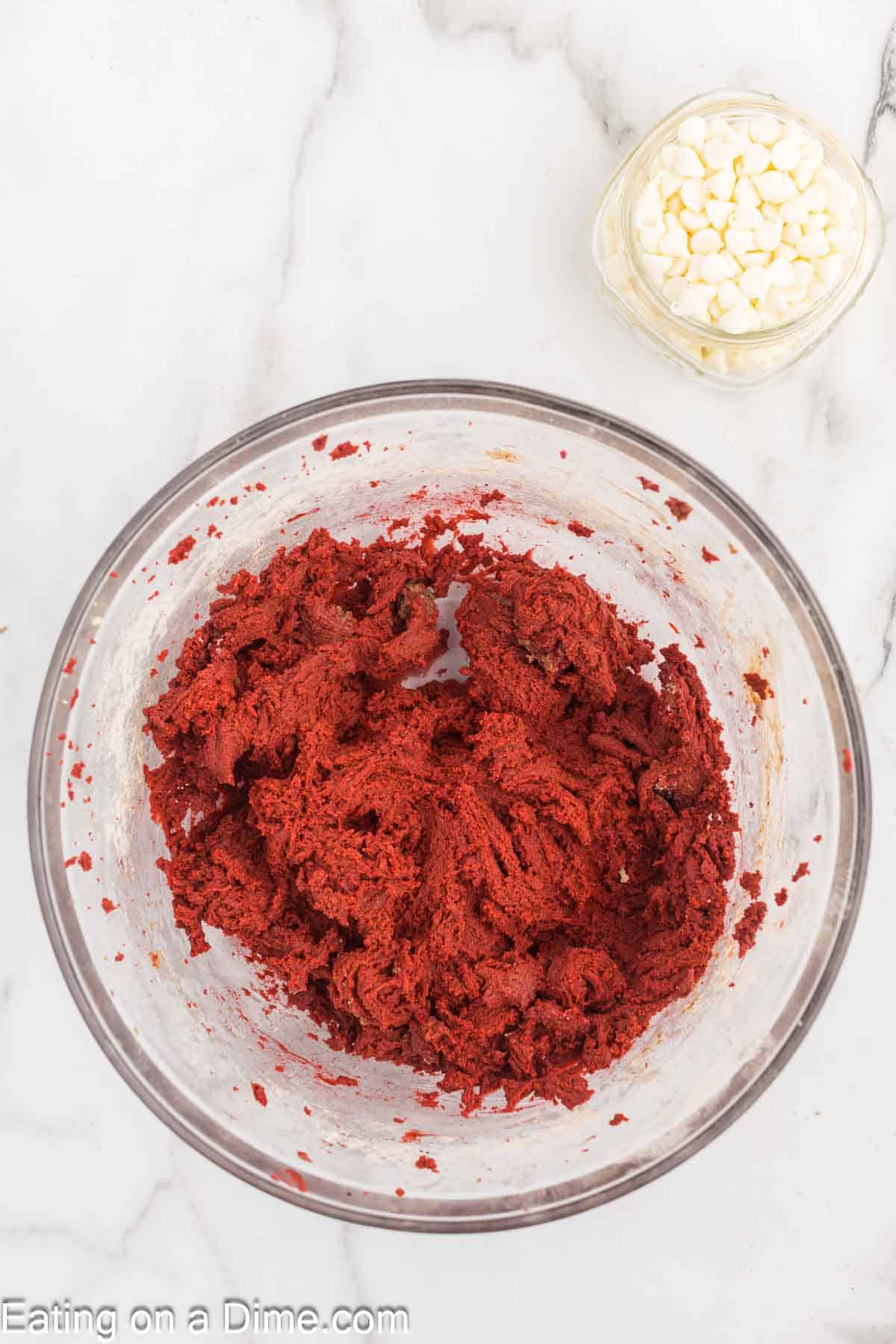 A glass mixing bowl filled with red velvet cookie dough sits on a white marble surface. A small glass bowl with white chocolate chips is placed beside the larger bowl. The lower left corner of the image has text that reads "Eating on a Dime.com.