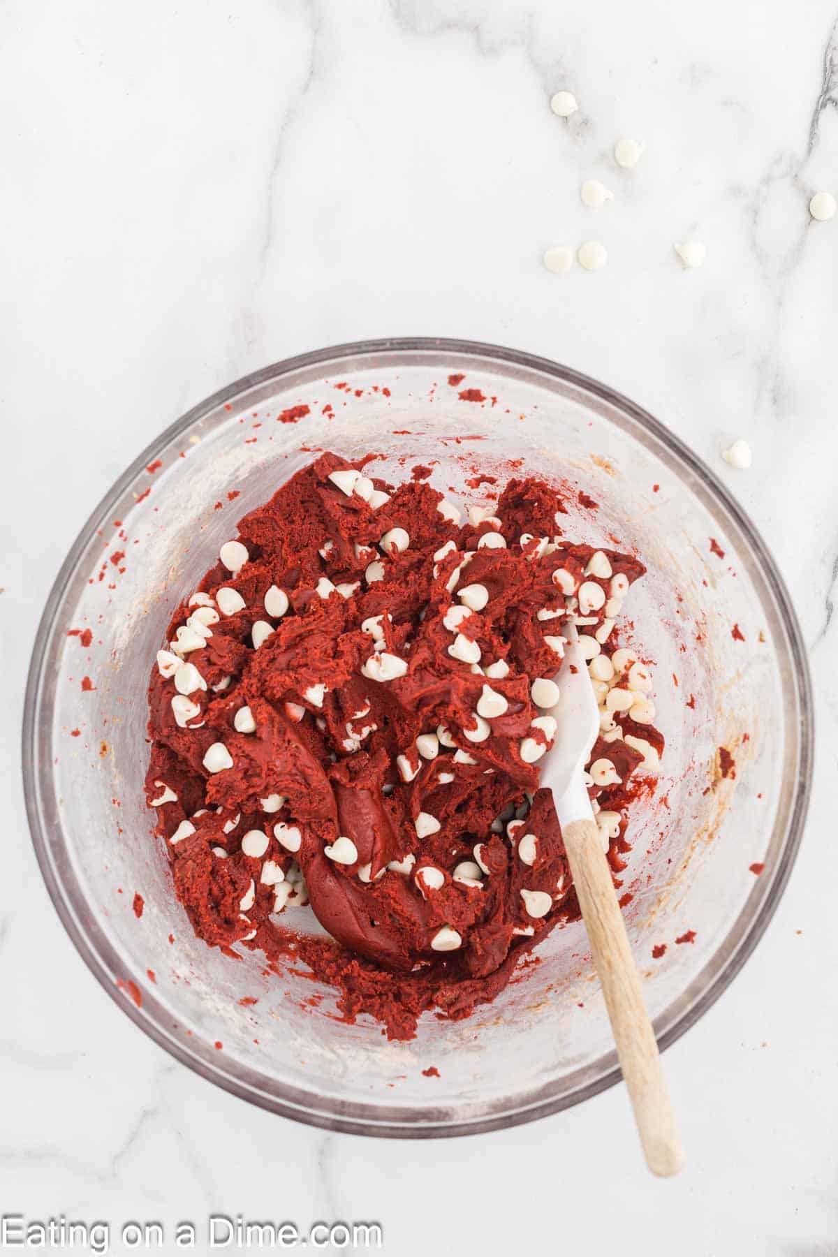 A clear glass bowl filled with red velvet cookie dough and white chocolate chips. A white spatula with a wooden handle is placed inside the bowl, and a few white chocolate chips are scattered on the white marble countertop nearby.