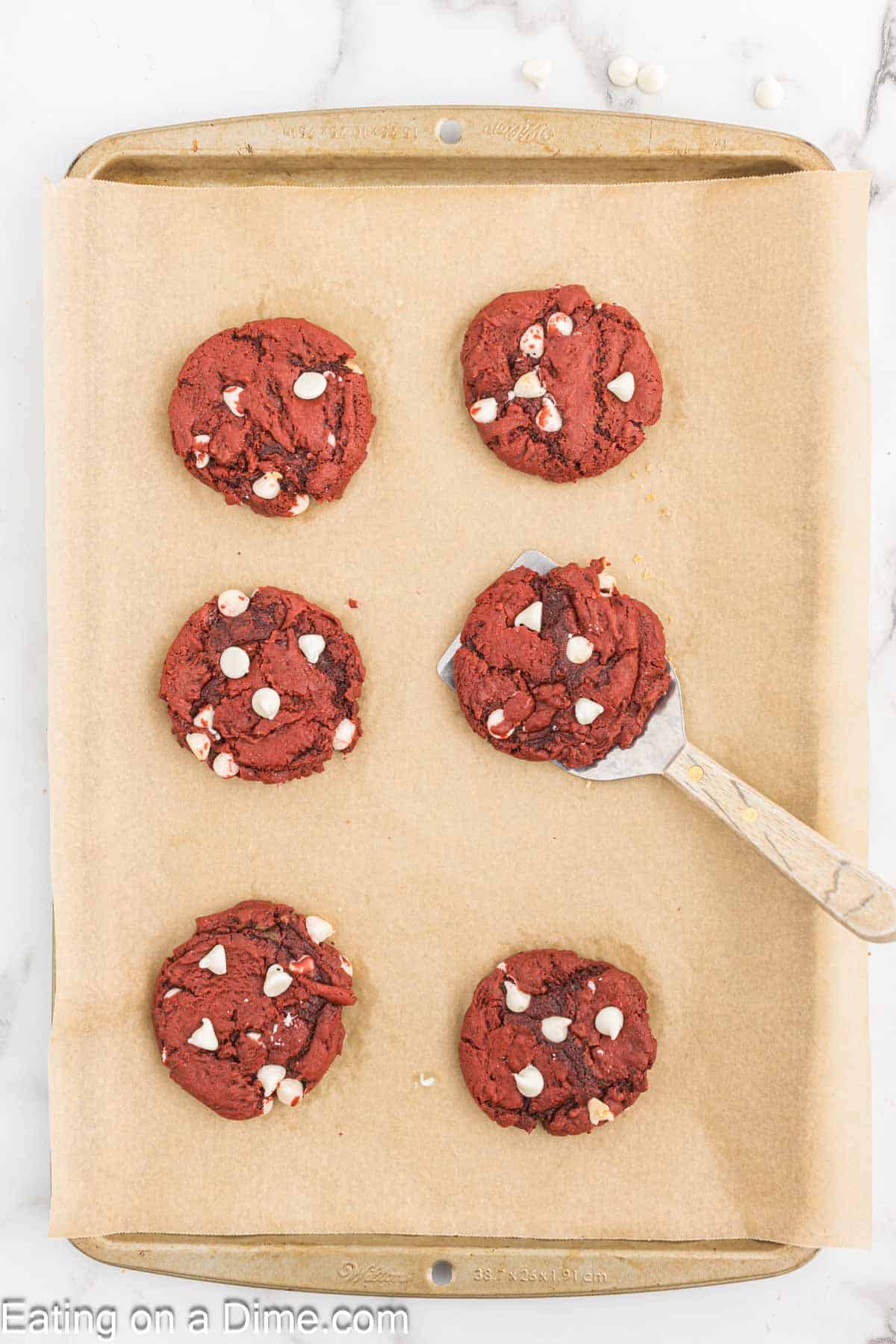 A baking sheet lined with parchment paper holds six Red Velvet Cookies, each sprinkled with white chocolate chips. A spatula is lifting one cookie, showing its underside. The baking sheet sits on a white marble surface.