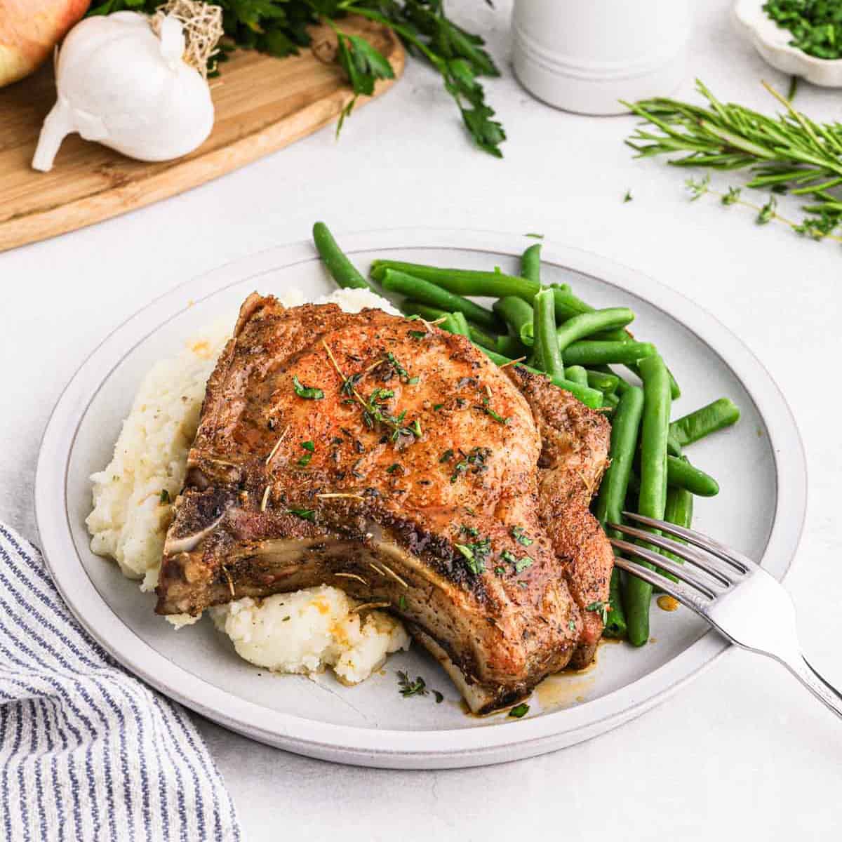 A plated meal consisting of a seasoned baked bone-in pork chop on a bed of mashed potatoes, accompanied by green beans. Fresh herbs can be seen sprinkled on top of the pork chop. In the background, fresh garlic, herbs, and a cutting board are visible. A fork rests on the plate.