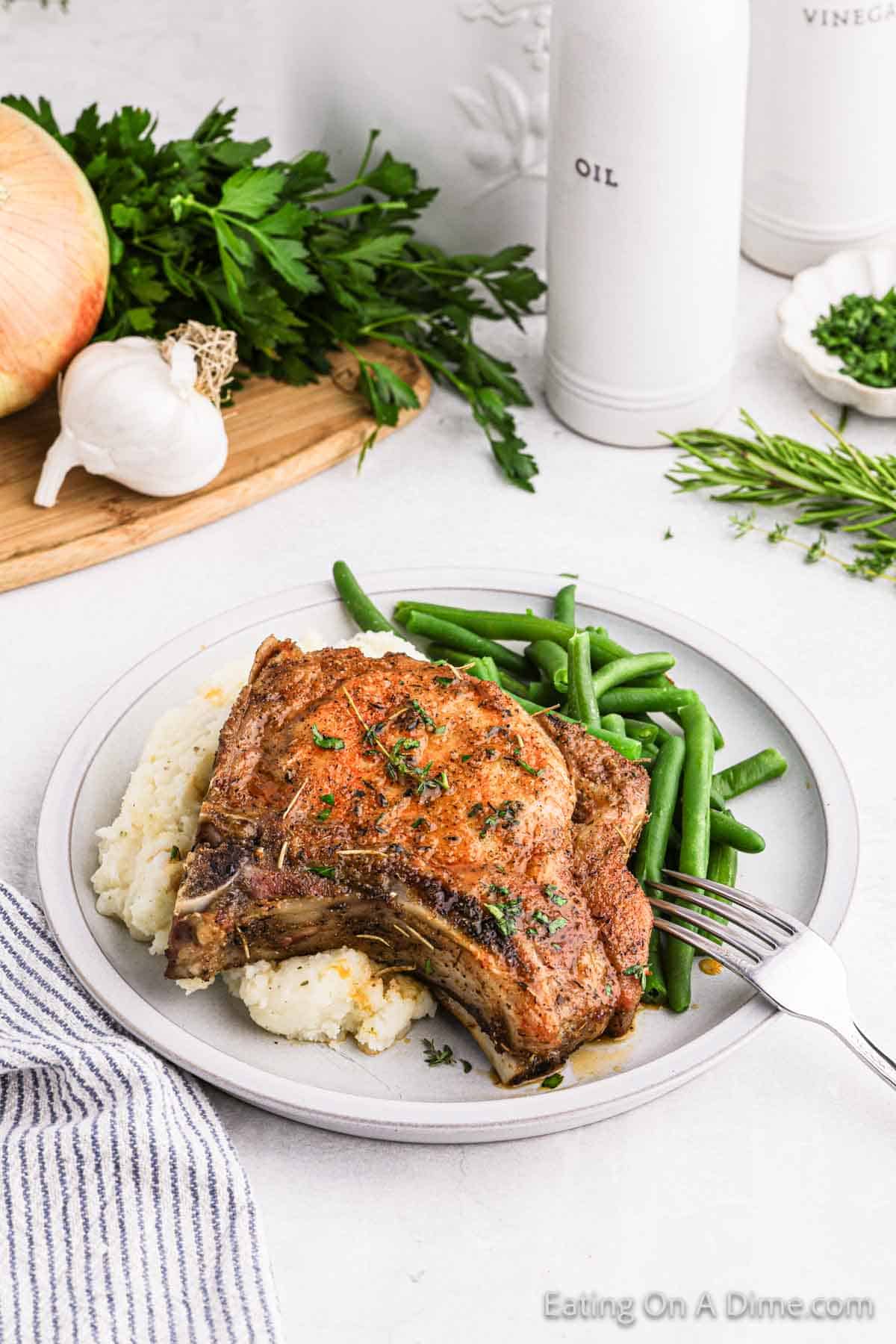 A plate featuring seasoned baked bone-in pork chops on a bed of mashed potatoes, accompanied by a serving of green beans. In the background, there are herbs, an onion, garlic, and bottles labeled OIL and VINEGAR. A fork rests on the plate's right side.