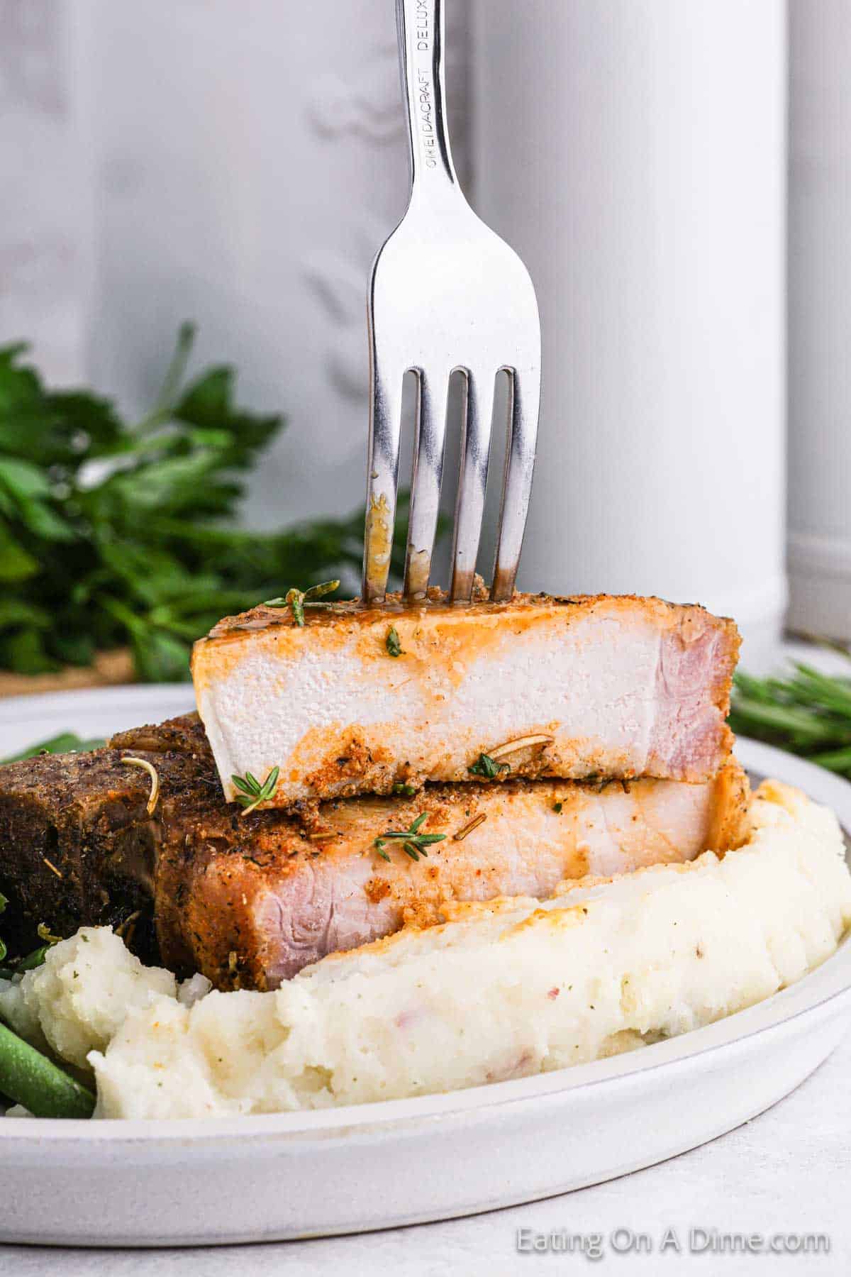 A close-up of a plate with two seasoned and cooked bone-in pork chops stacked on top of each other, garnished with herbs. The dish is served with mashed potatoes and a side of green beans. A fork is inserted into the top pork chop. A bunch of fresh parsley is in the background.