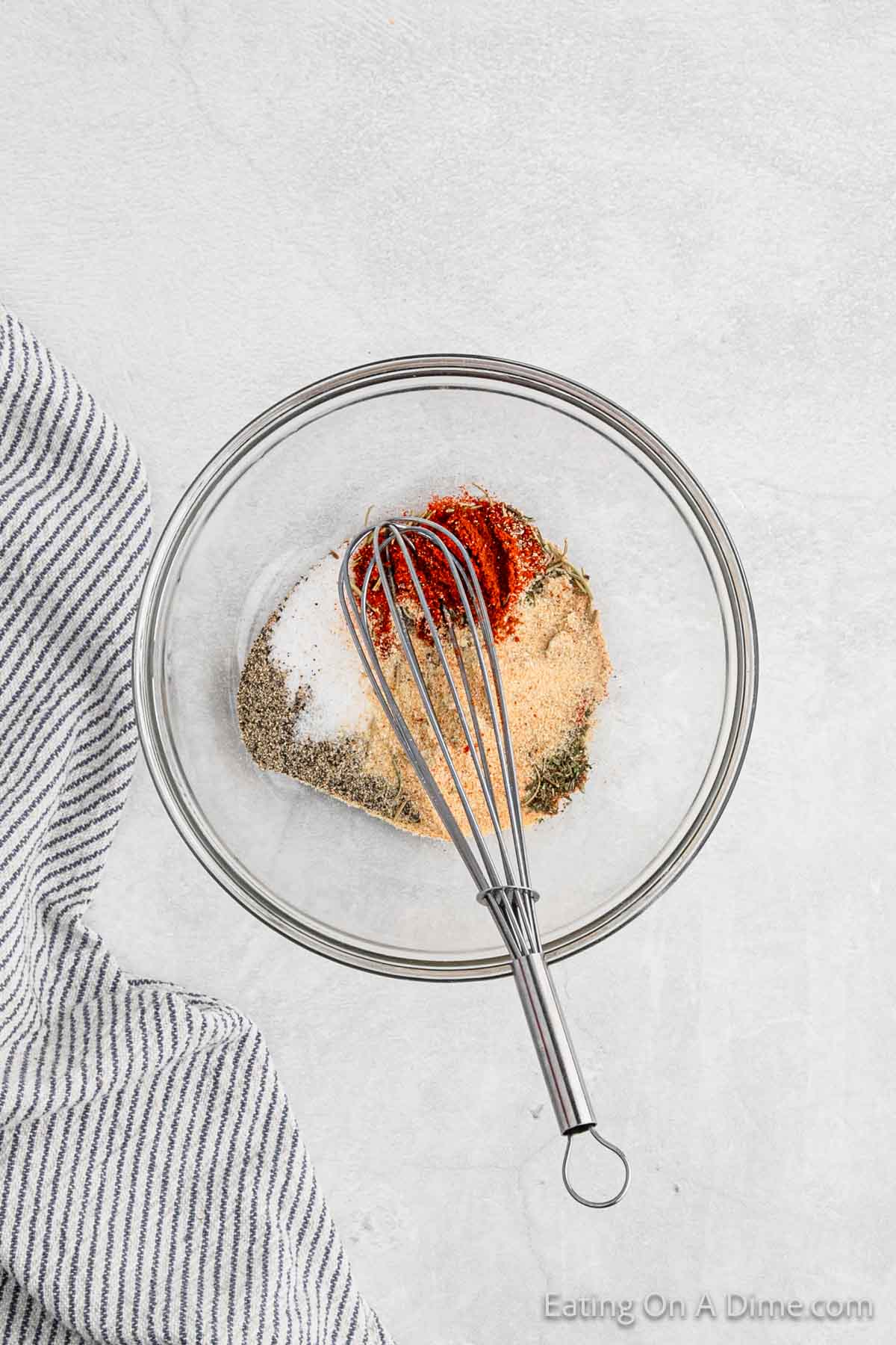 A glass bowl filled with various spices, including paprika, garlic powder, salt, pepper, and other herbs sits ready for seasoning Baked Pork Chops. A metal whisk is partially submerged in the mixture. A striped cloth napkin rests to the side on a light gray surface.