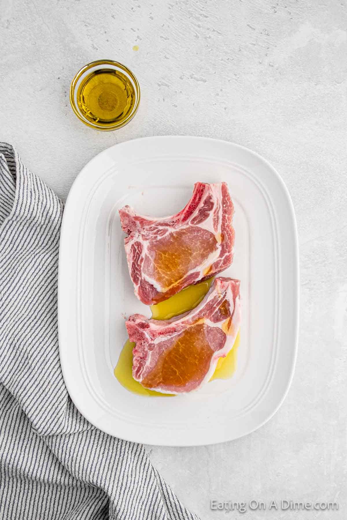 Two raw baked bone-in pork chops marinating in olive oil on a white rectangular plate. A small clear bowl filled with olive oil is beside the plate on a light gray surface. A black and white striped cloth napkin is partially visible to the left of the plate.