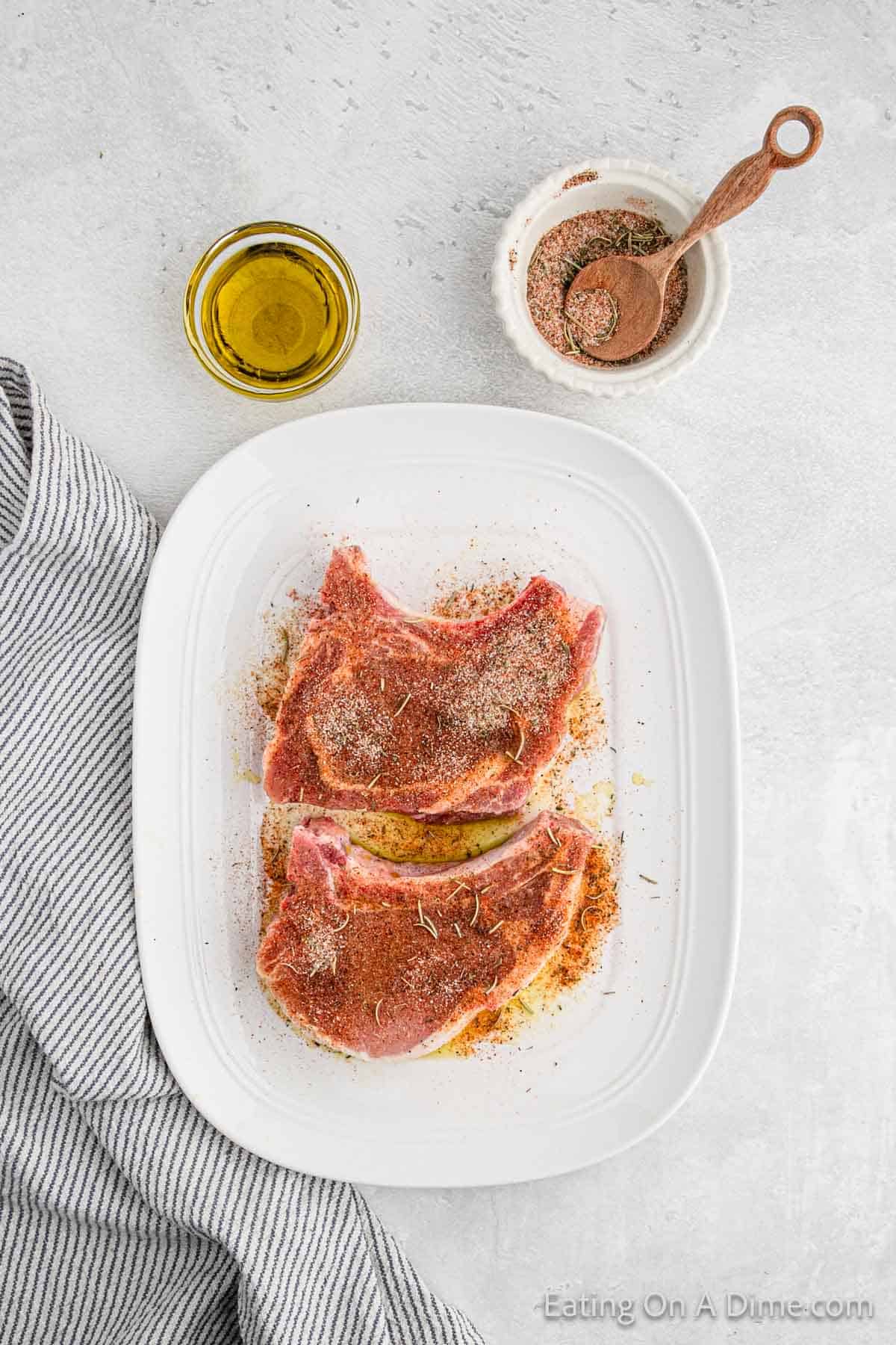 A rectangular white dish contains two raw, seasoned bone-in pork chops. To the side of the dish is a small bowl of seasoning with a wooden spoon resting on it. A small, round glass container of olive oil sits beside a blue and white striped cloth.