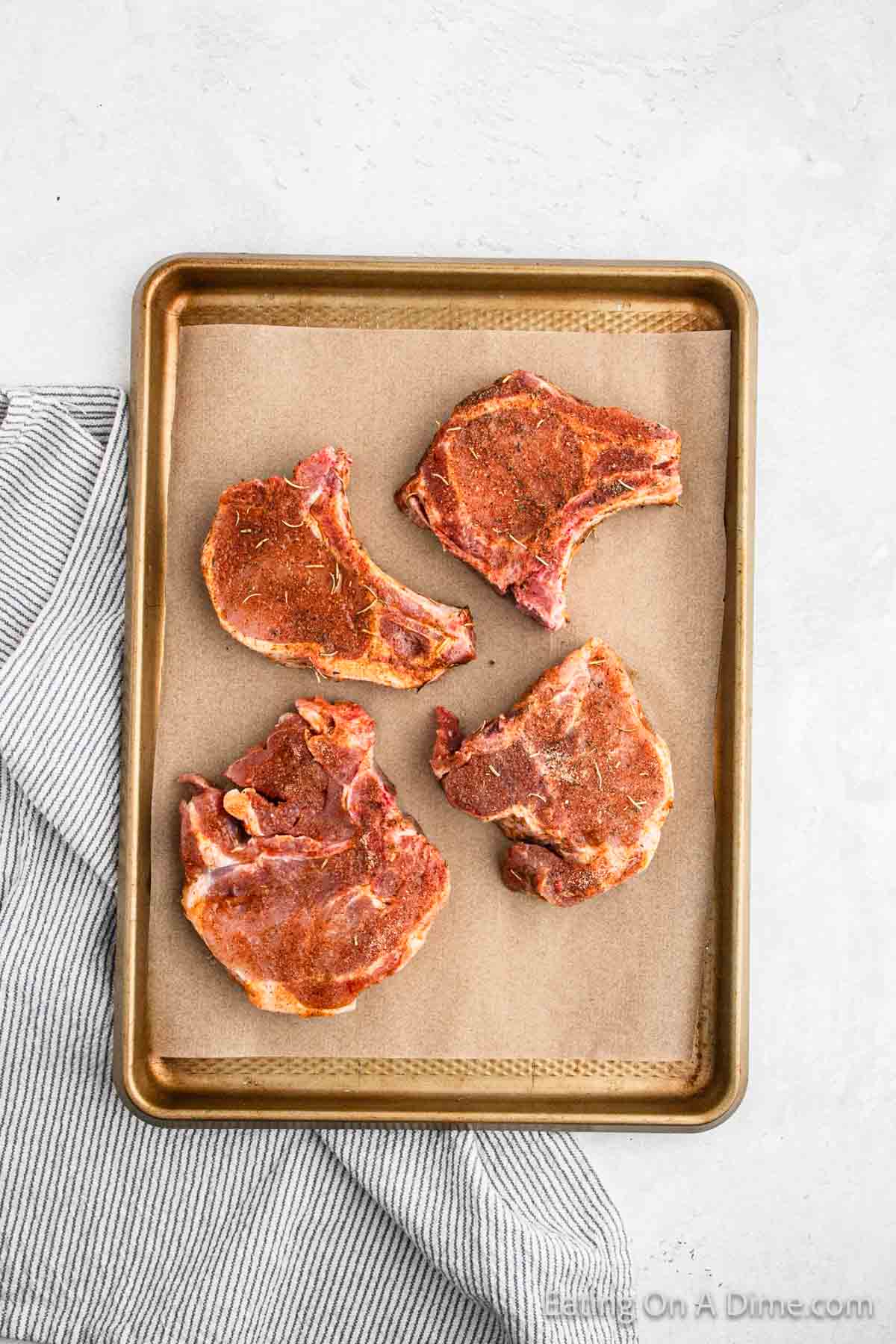 A baking sheet with four seasoned bone-in pork chops arranged on parchment paper. The baking sheet is placed on a light gray surface, with part of a gray and white striped kitchen towel visible in the bottom left corner, ready to be baked to perfection.