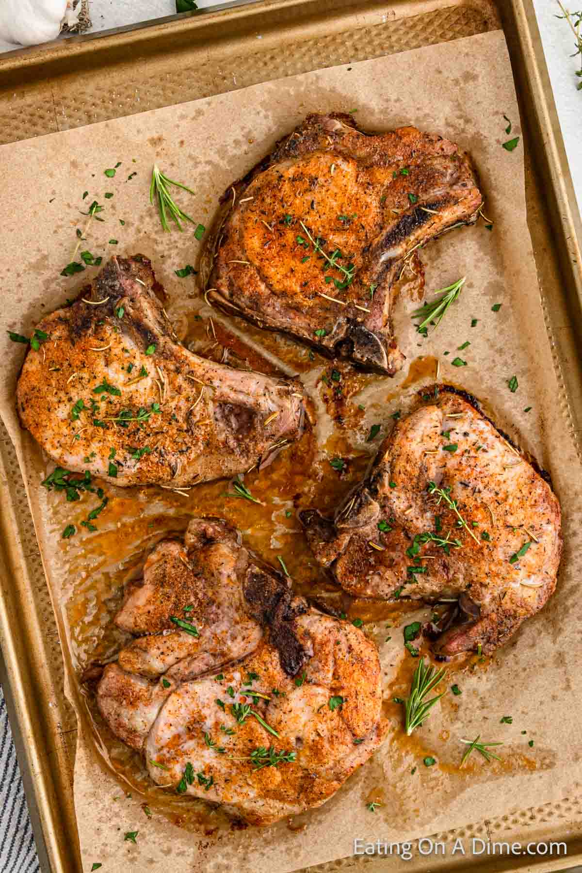 Top view of a baking sheet with four bone-in pork chops, garnished with fresh herbs. The brown, crispy edges and juices around the chops indicate they were oven baked. The image is from the food blog "Eating On A Dime.