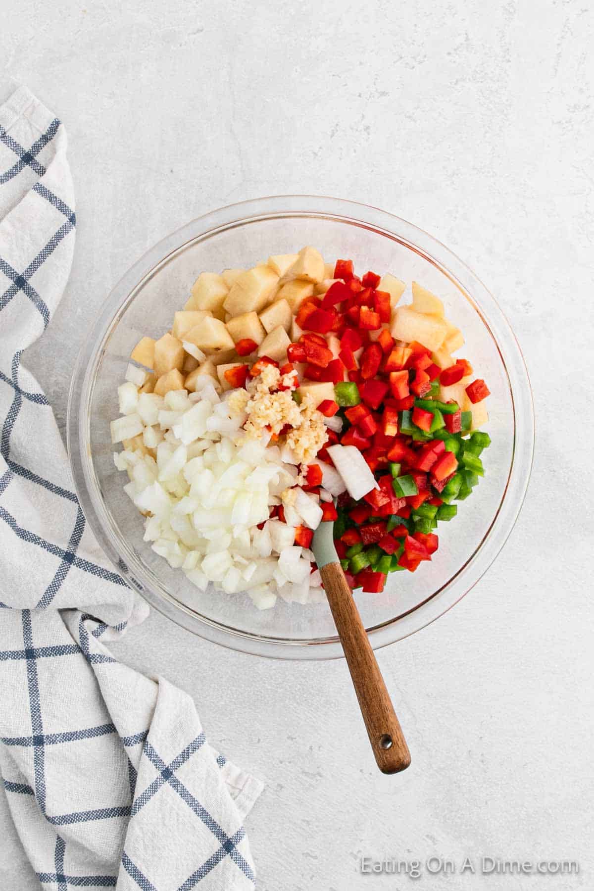 A glass bowl filled with chopped onions, red bell peppers, green bell peppers, cubed potatoes, and minced garlic is ready for a breakfast recipe. A wooden spoon is placed in the bowl. A white and blue checkered towel is partially visible to the left of the bowl on a light countertop.