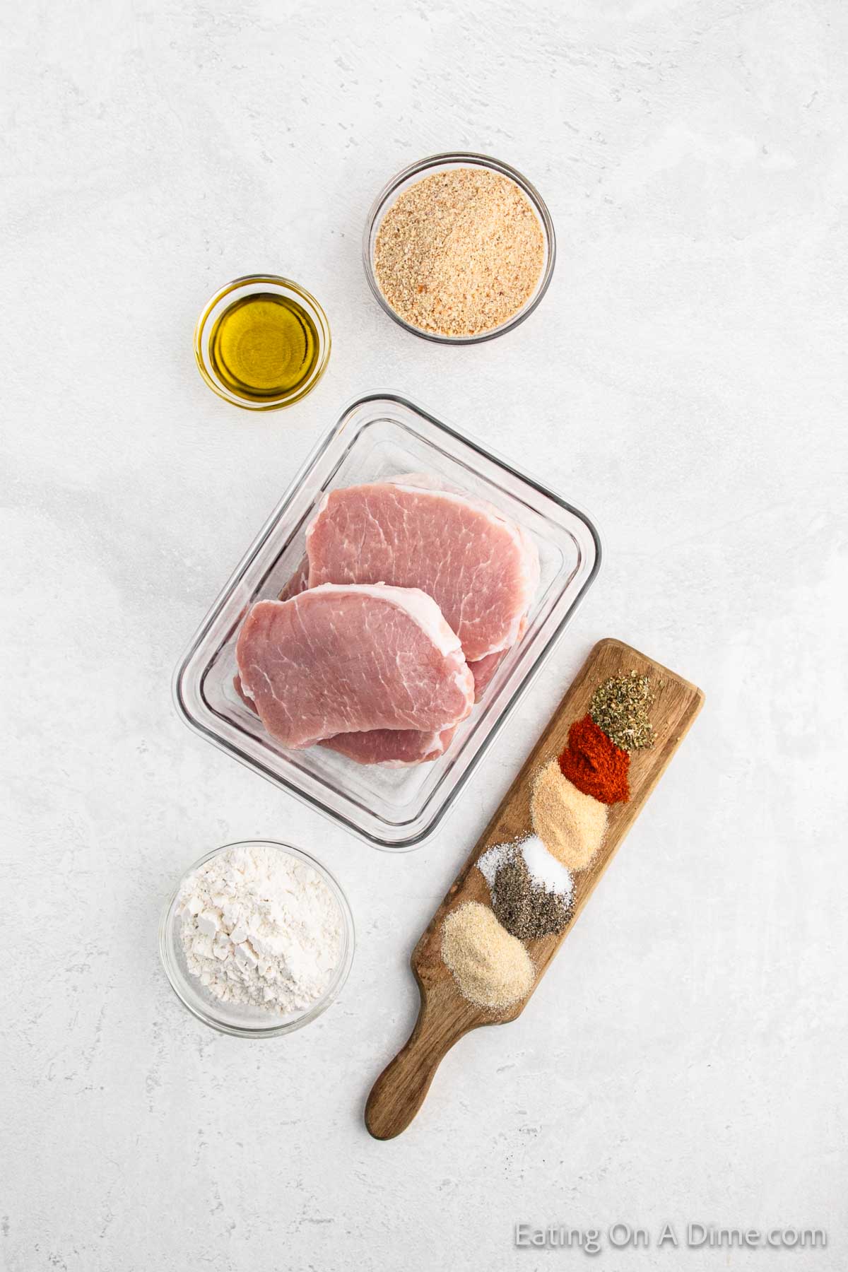 A flat lay of ingredients for a homemade recipe. At the center is a glass container with raw pork chops, perfect for a Shake and Bake meal. Surrounding it are small bowls of breadcrumbs, olive oil, and flour. A wooden board holds various spices, including paprika, black pepper, and garlic powder.