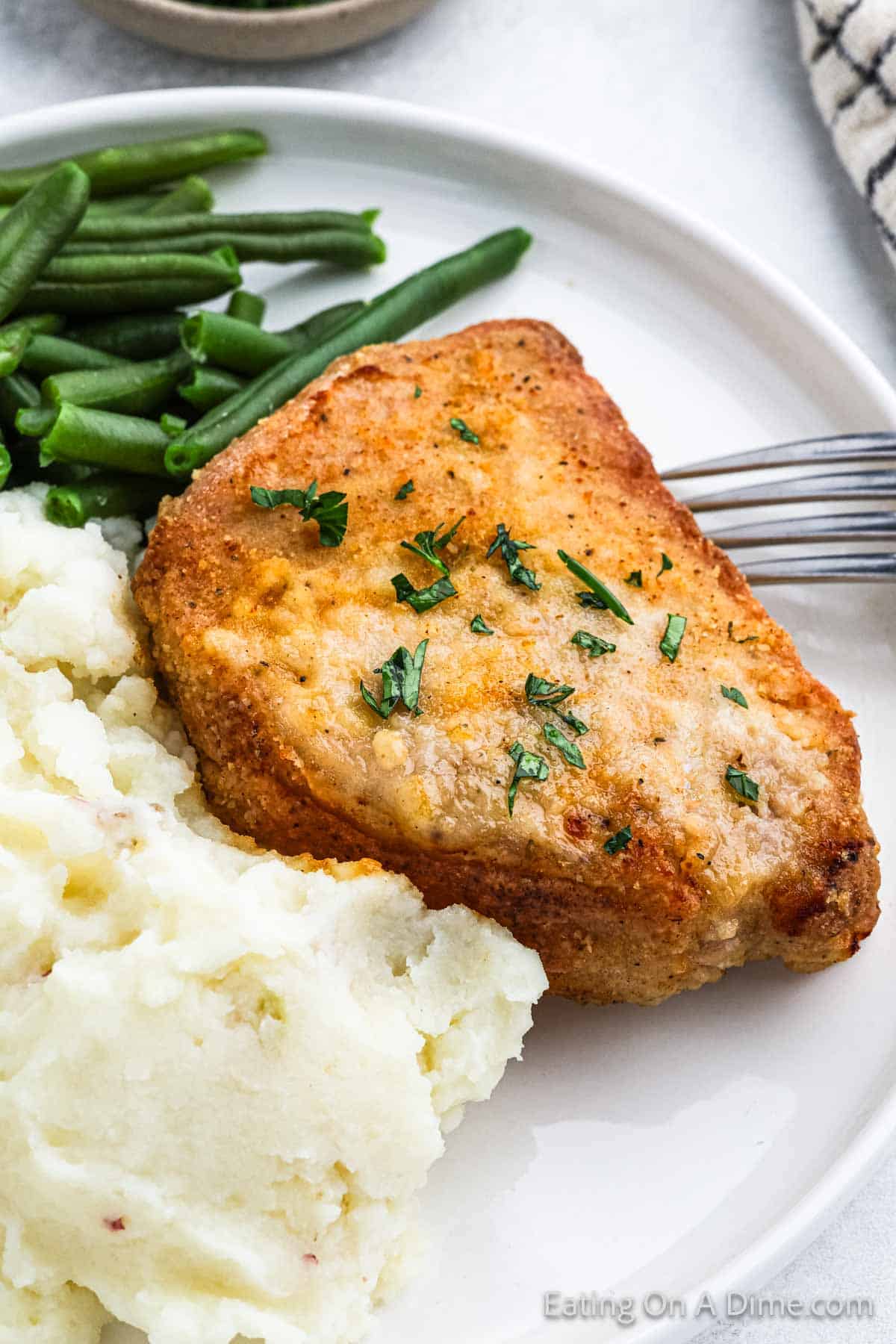 A plate featuring a golden-brown, Shake and Bake seasoned pork chop garnished with chopped herbs, accompanied by a serving of mashed potatoes and green beans. A fork is placed on the plate next to the homemade pork chop. The background includes a textured cloth napkin.