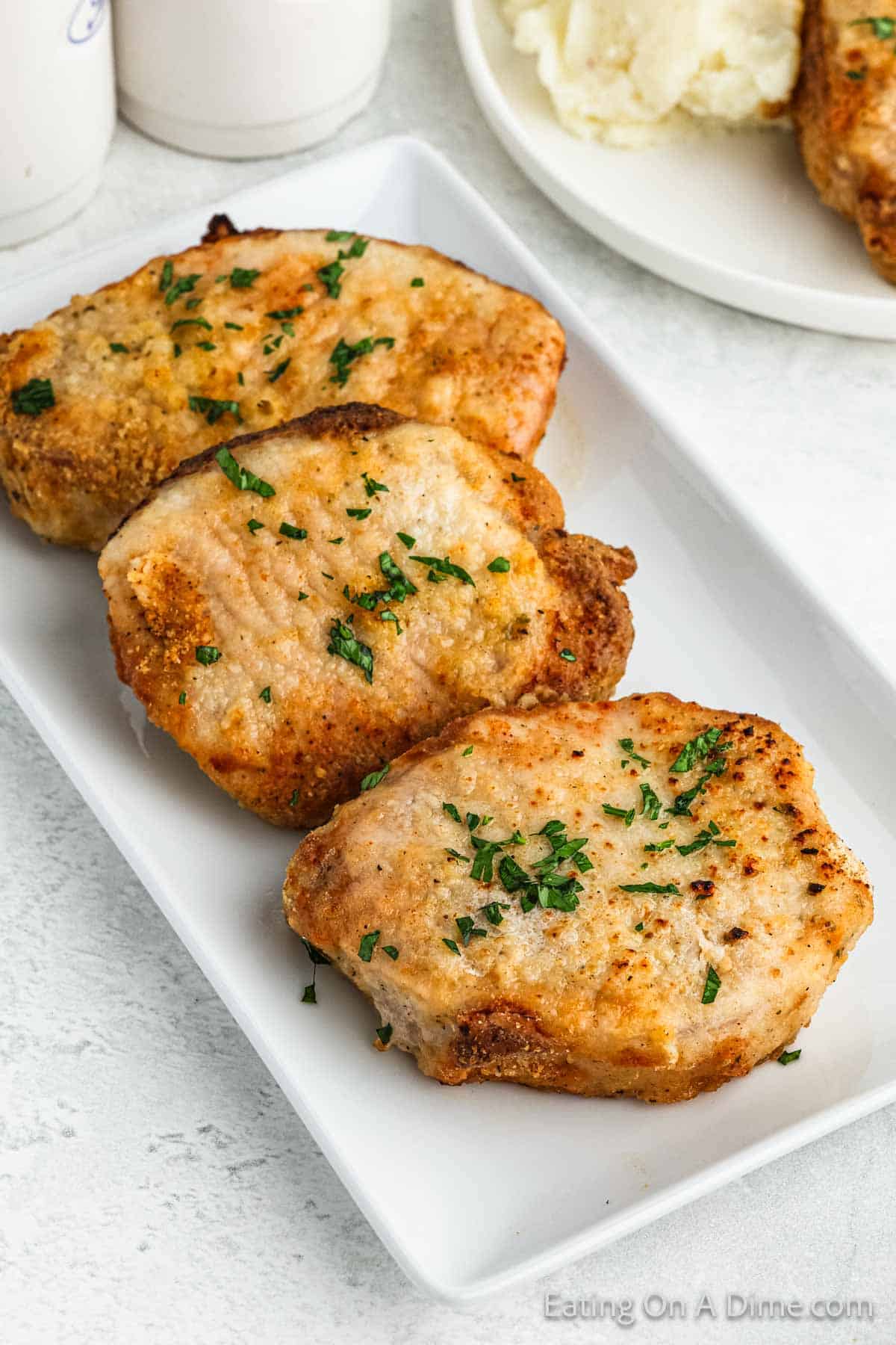 A white rectangular plate holds three seasoned, golden-brown Shake and Bake pork chops garnished with chopped parsley. In the background, there is a white plate with homemade mashed potatoes and another pork chop, along with partial views of salt and pepper shakers.