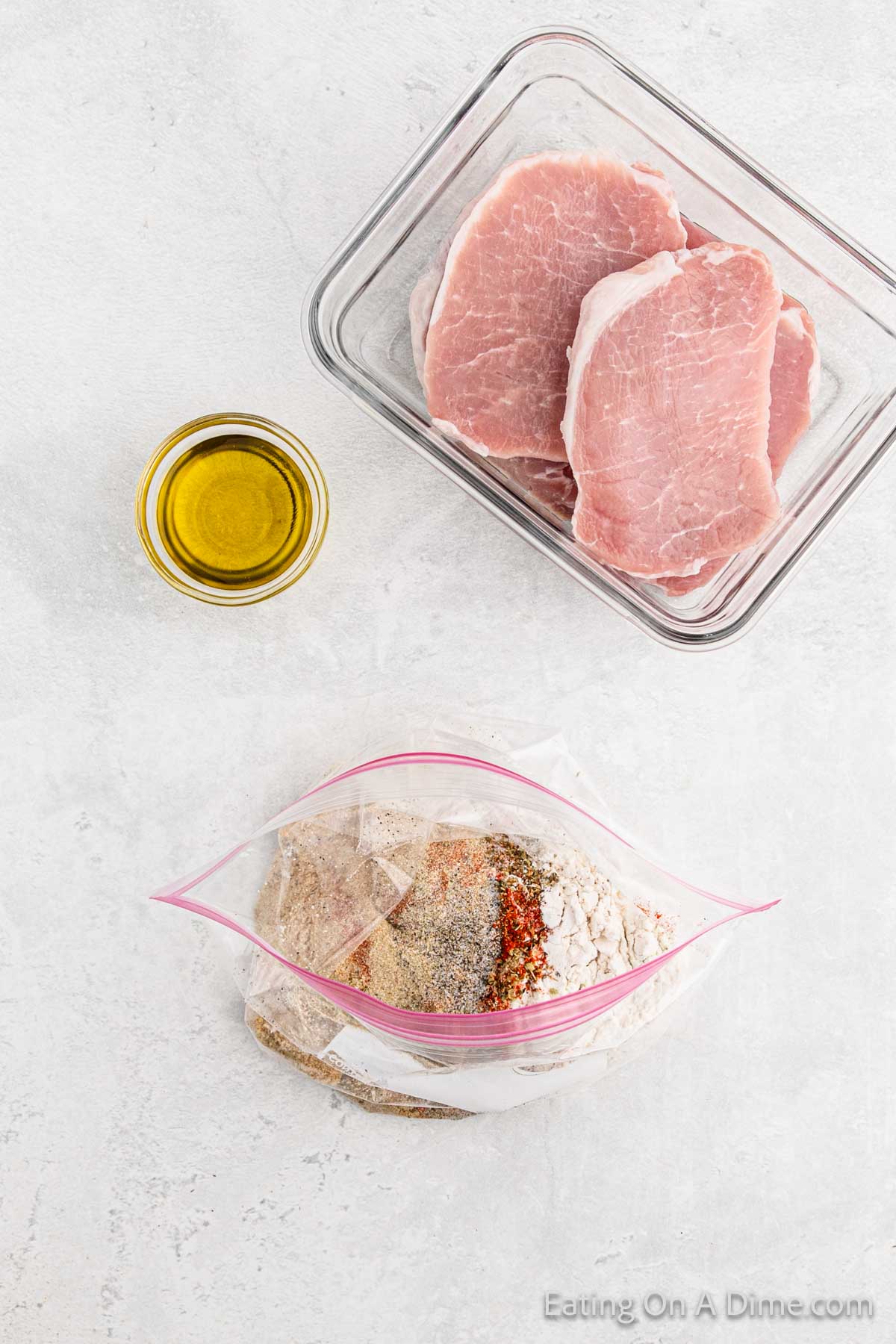 A glass container holding raw pork chops, a small glass bowl filled with olive oil, and an open plastic zip-lock bag with flour and various spices for a homemade Shake and Bake are laid out on a white countertop.