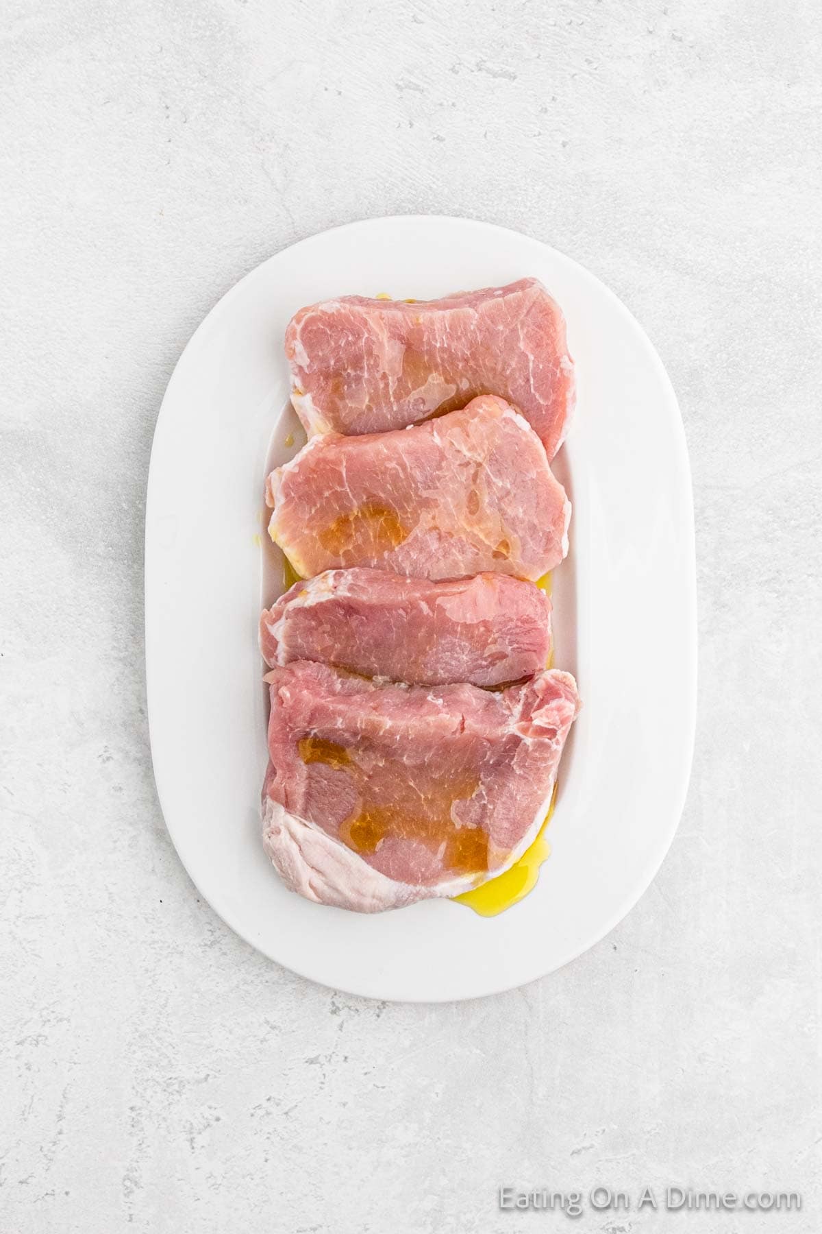 Four raw homemade pork chops are arranged on a white oval plate, coated with oil. The plate is set on a light gray textured surface.