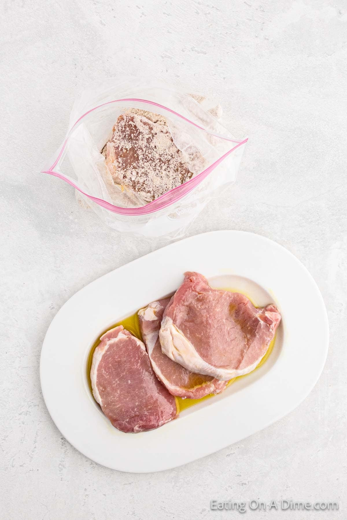 Homemade pork chops on a white plate with olive oil next to a plastic bag containing a Shake and Bake-seasoned pork chop coated in crushed pork rinds. The background is a white, textured surface.