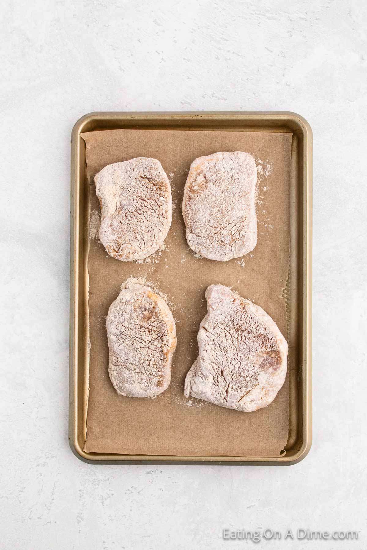 Four pieces of raw, breaded chicken are placed on a parchment-lined baking sheet. The light breading reminiscent of a homemade Shake and Bake recipe is visible on the chicken pieces, which are evenly spaced out. The baking sheet is set against a plain, light-colored background.