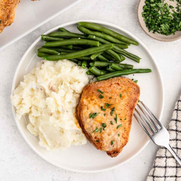 A plate of food featuring a seasoned Shake and Bake pork chop garnished with herbs, a serving of mashed potatoes, and a portion of green beans. A fork is placed on the right side of the plate. In the background, there's a small bowl of chopped parsley and a striped cloth napkin.