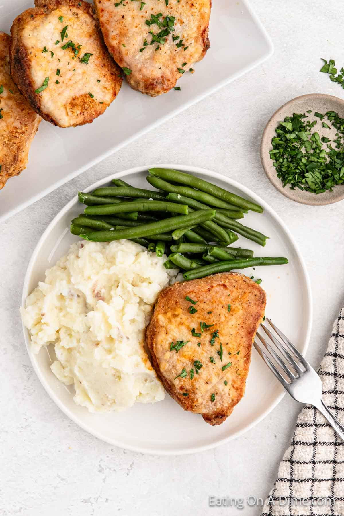 A plate with a meal featuring a perfectly cooked Shake and Bake pork chop garnished with herbs, accompanied by creamy mashed potatoes and green beans. A fork rests on the plate. In the background, there is a dish with additional homemade pork chops and a small bowl of chopped herbs.