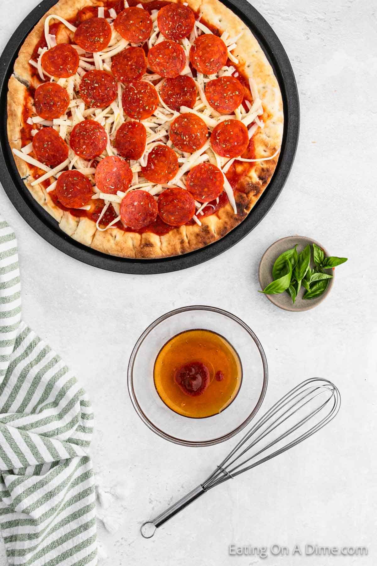 Slice pepperoni, mozzarella cheese and pizza sauce on pizza crust with a small bowl of honey and basil with a whisk