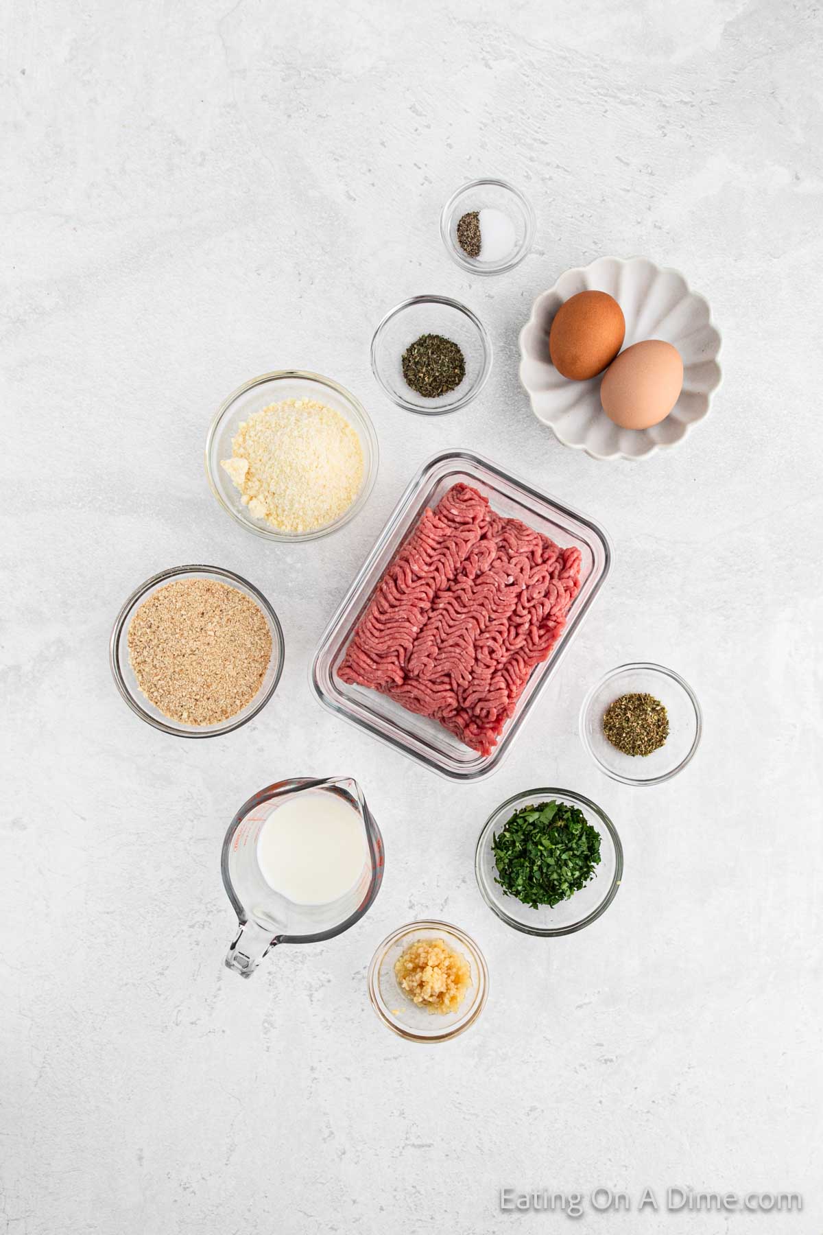 Top view of a variety of small bowls filled with ingredients for Italian meatballs. From left to right, they contain breadcrumbs, parmesan, dried herbs, minced garlic, chopped parsley, black pepper, olive oil, milk, and two brown eggs. A container of ground meat is centered.